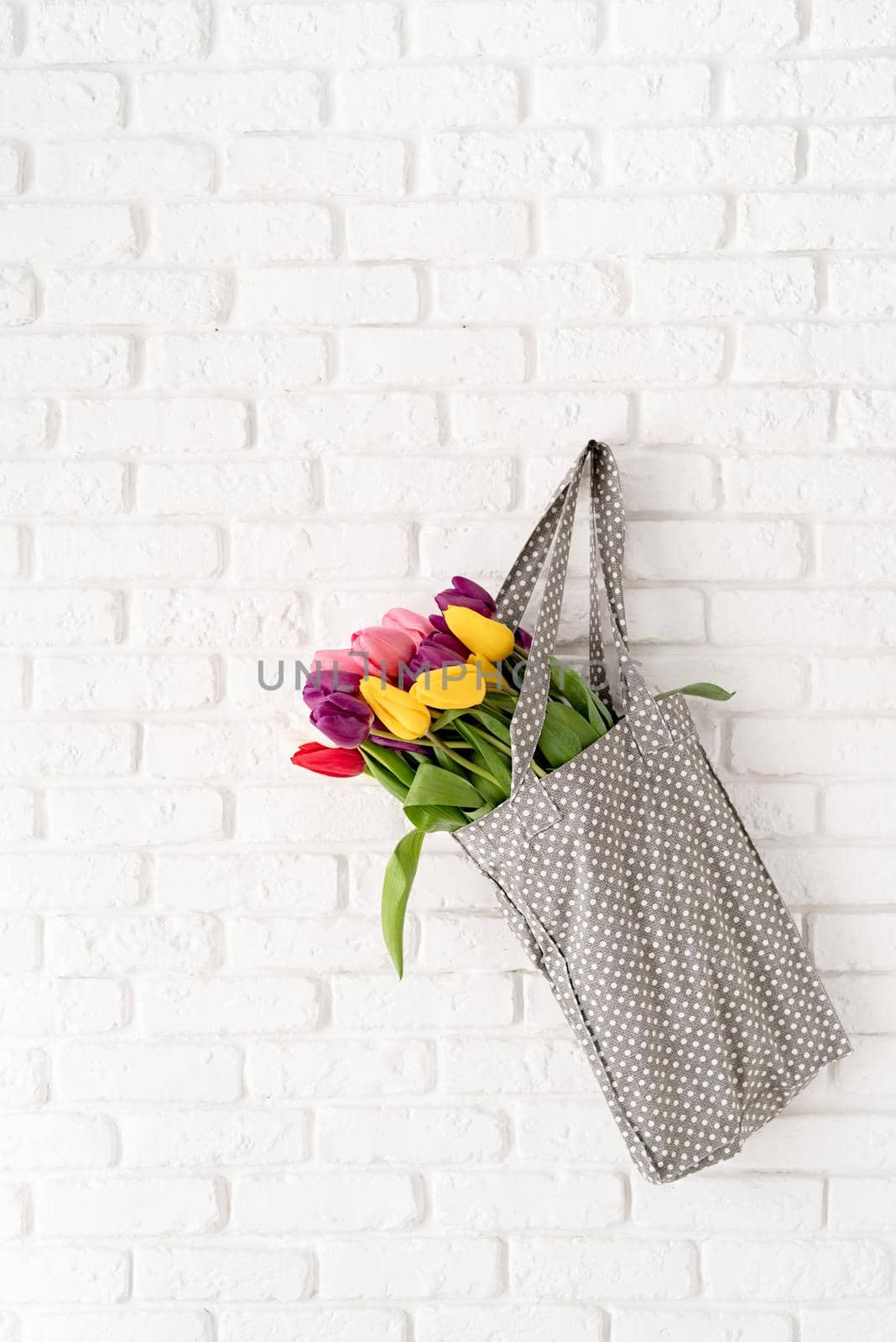 Eco friendly, zero waste concept. Gray polka dot fabric bag full of colorful tulips on white brick background