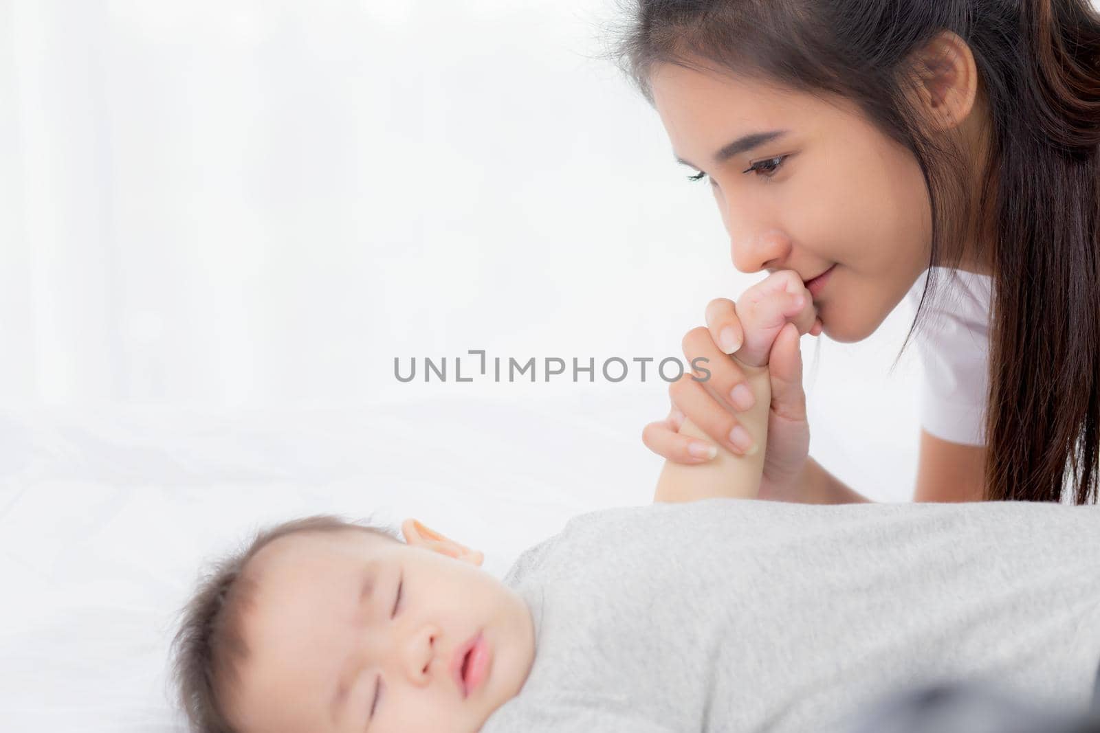 Young asian mother kiss hand of little baby girl with tender on bed in the bedroom, mom love newborn and care, mother with expression with child together, parent and daughter, family concept.