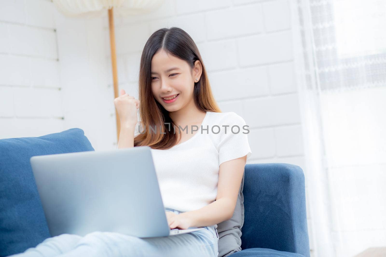 Young asian business woman smile and work from home with laptop computer online to internet on sofa in living room, freelance girl using notebook on couch with comfort, new normal, lifestyle concept.