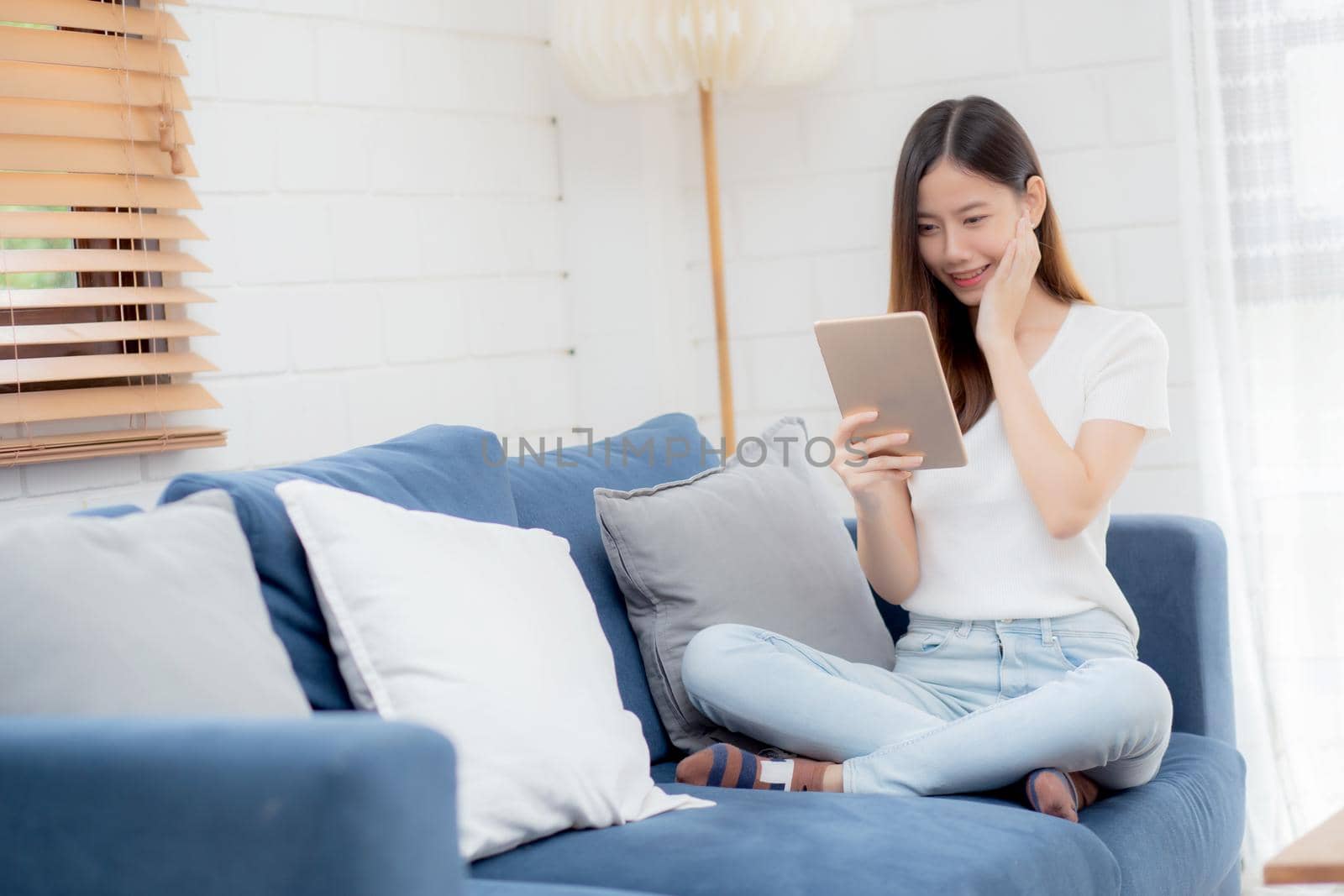 Young attractive asian woman resting using browsing tablet computer on sofa at home, happy girl sitting on couch relax reading digital gadget with excited, communication and lifestyle concept.