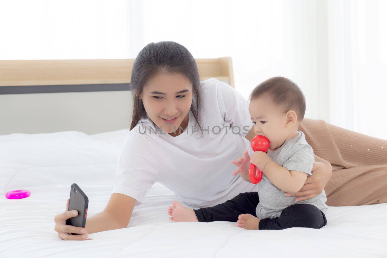 Young asian mother and little baby girl or newborn selfie with smart phone on bed in bedroom, happiness mom and daughter using phone video call at home, two people, family and communication concept. by nnudoo