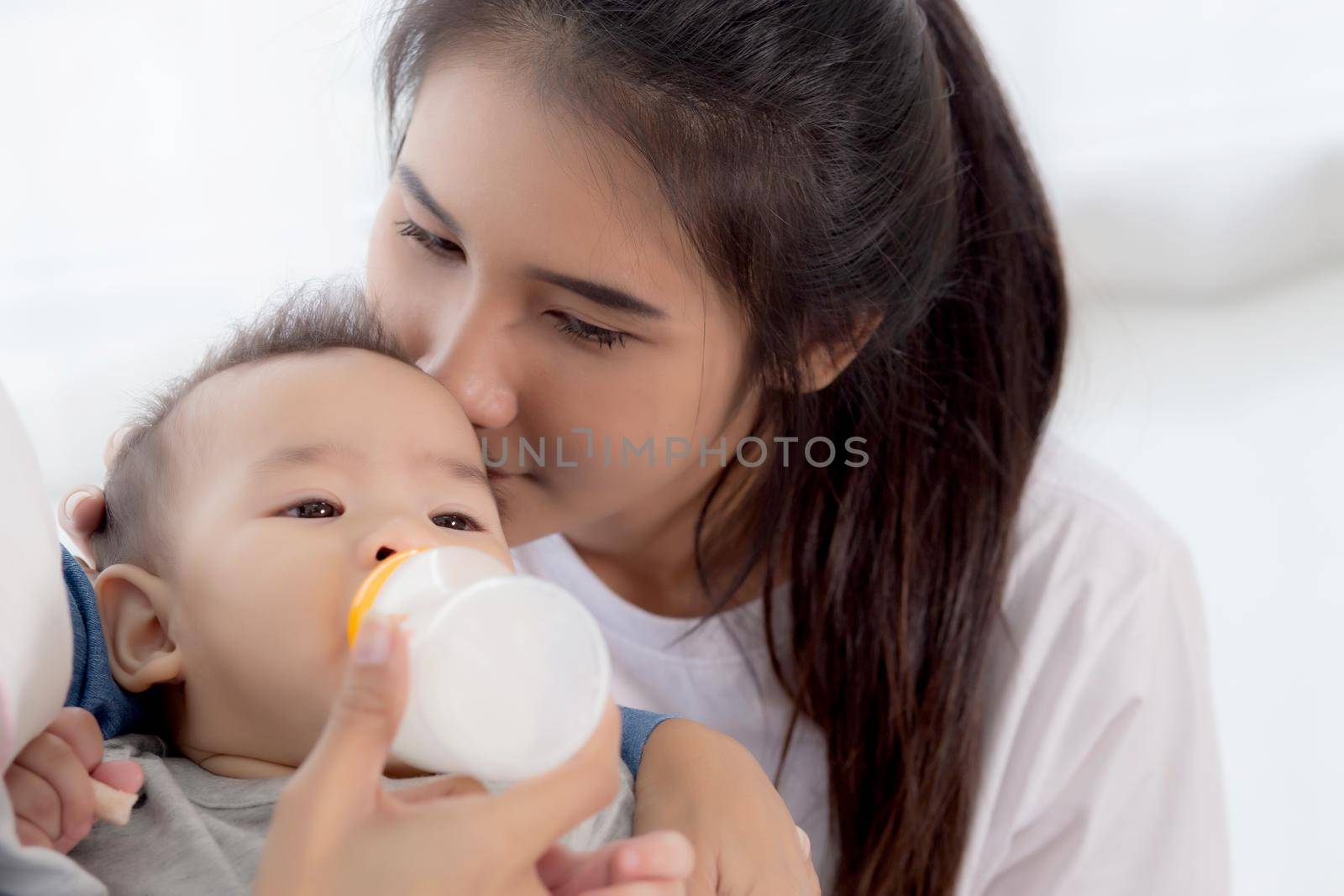 Young asian mother embracing and feeding little baby girl with bottle of milk at home, newborn innocence drinking with mom satisfied, relationship and bonding of mum and child, family concept.