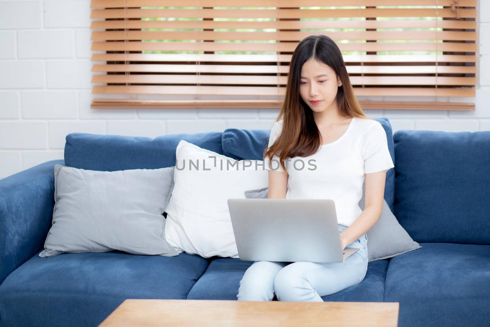 Young asian business woman smile and work from home with laptop computer online to internet on sofa in living room, freelance girl using notebook on couch with comfort, new normal, lifestyle concept.