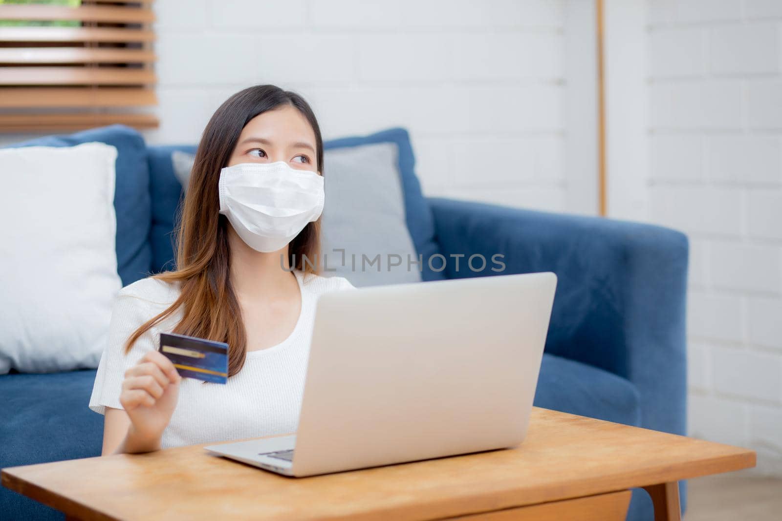 Young asian woman in face mask holding credit card shopping online with laptop computer buy and payment, girl using debit card purchase and thinking planning finance, lifestyle and new normal concept.