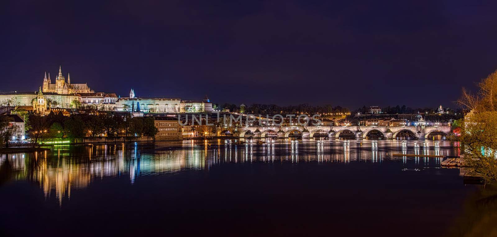 Charles Bridge in Prague in the evening with colorful lights from lanterns. In the river the reflection of the evening illuminations. Panorama