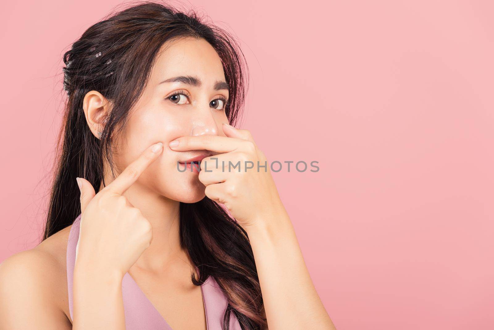 Portrait teenage Asian beautiful young woman having skin problems squeezing pimples on her face, studio shot on pink background, with copy space, Thai female acne, beauty care concept