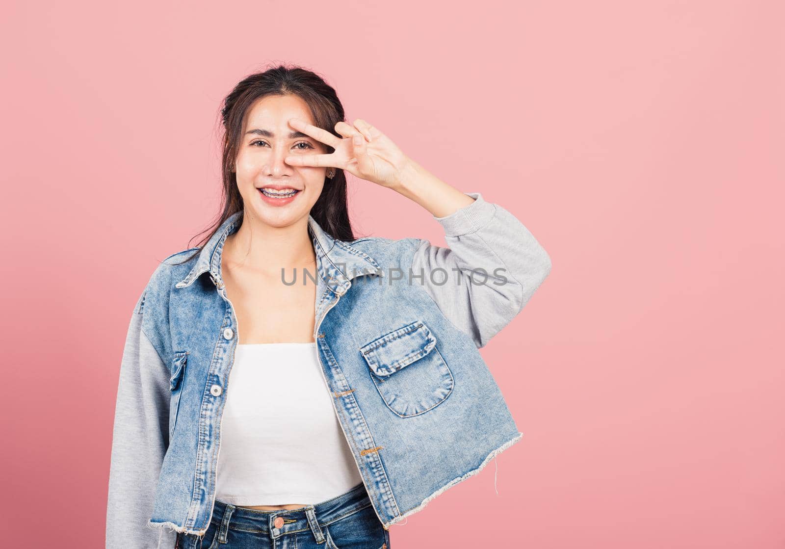 Asian happy portrait beautiful cute young woman wear denim smile standing showing finger making v-sign victory symbol near eye looking to camera studio shot isolated on pink background with copy space