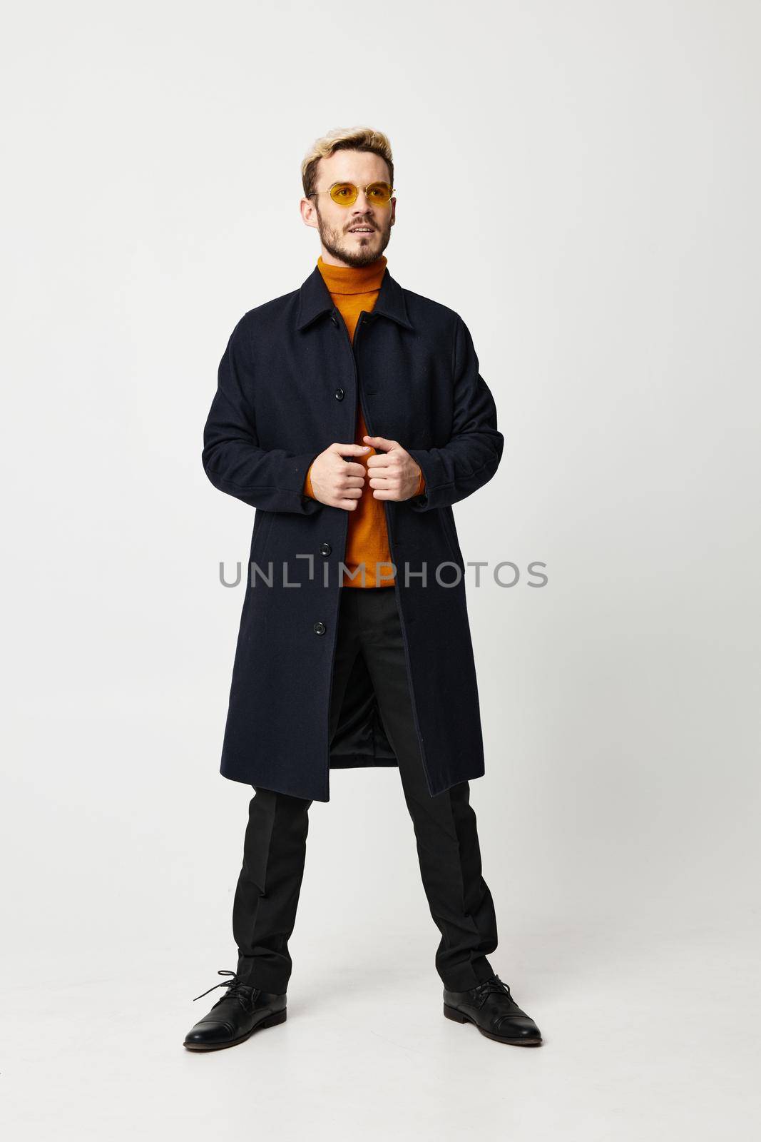 fashionable man in a dark coat spread his legs shoulder-width apart on a light background and an orange sweater model by SHOTPRIME
