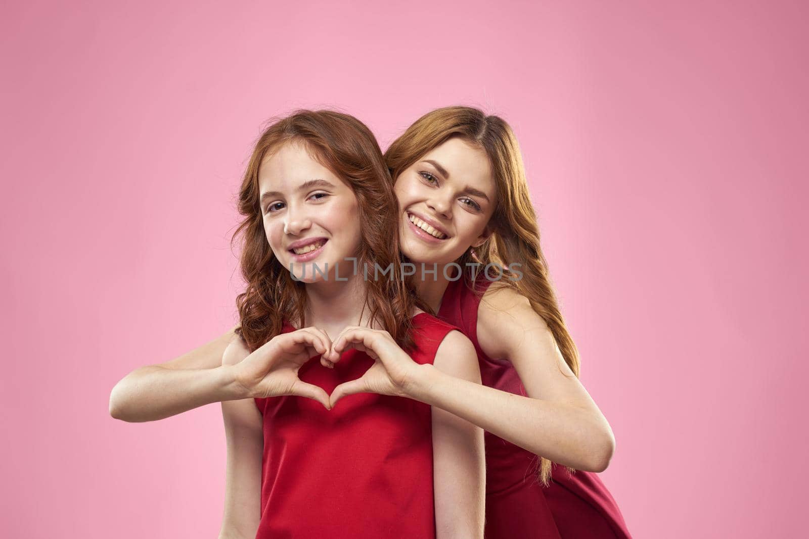 Cheerful mom and daughter wearing red dresses are standing next to joy family on pink background by SHOTPRIME