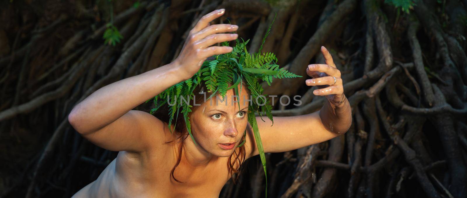 A beautiful young nude woman with fern wreath on her head enjoying nature in the forest river