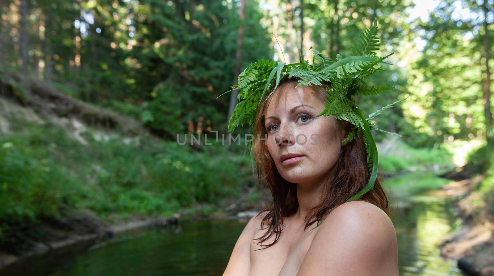 A beautiful young nude woman with fern wreath on her head enjoying nature in the forest river