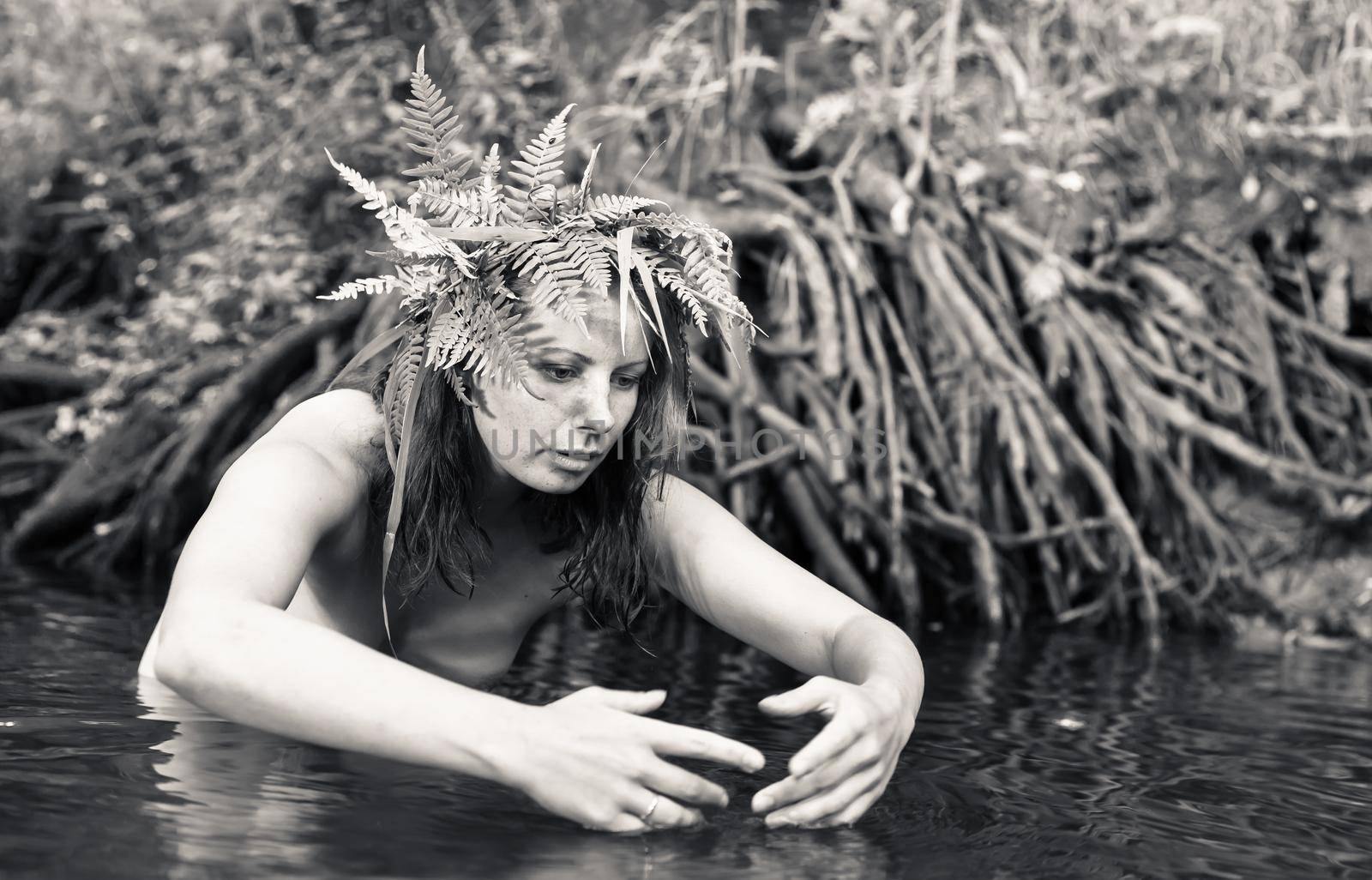 A beautiful young nude woman with fern wreath on her head enjoying nature in the forest river