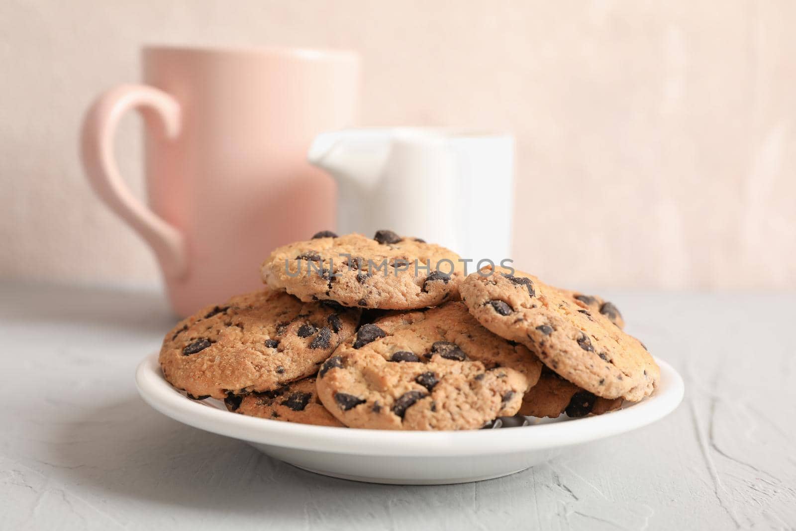 Plate with tasty chocolate chip cookies and blurred cup of milk on gray background, closeup