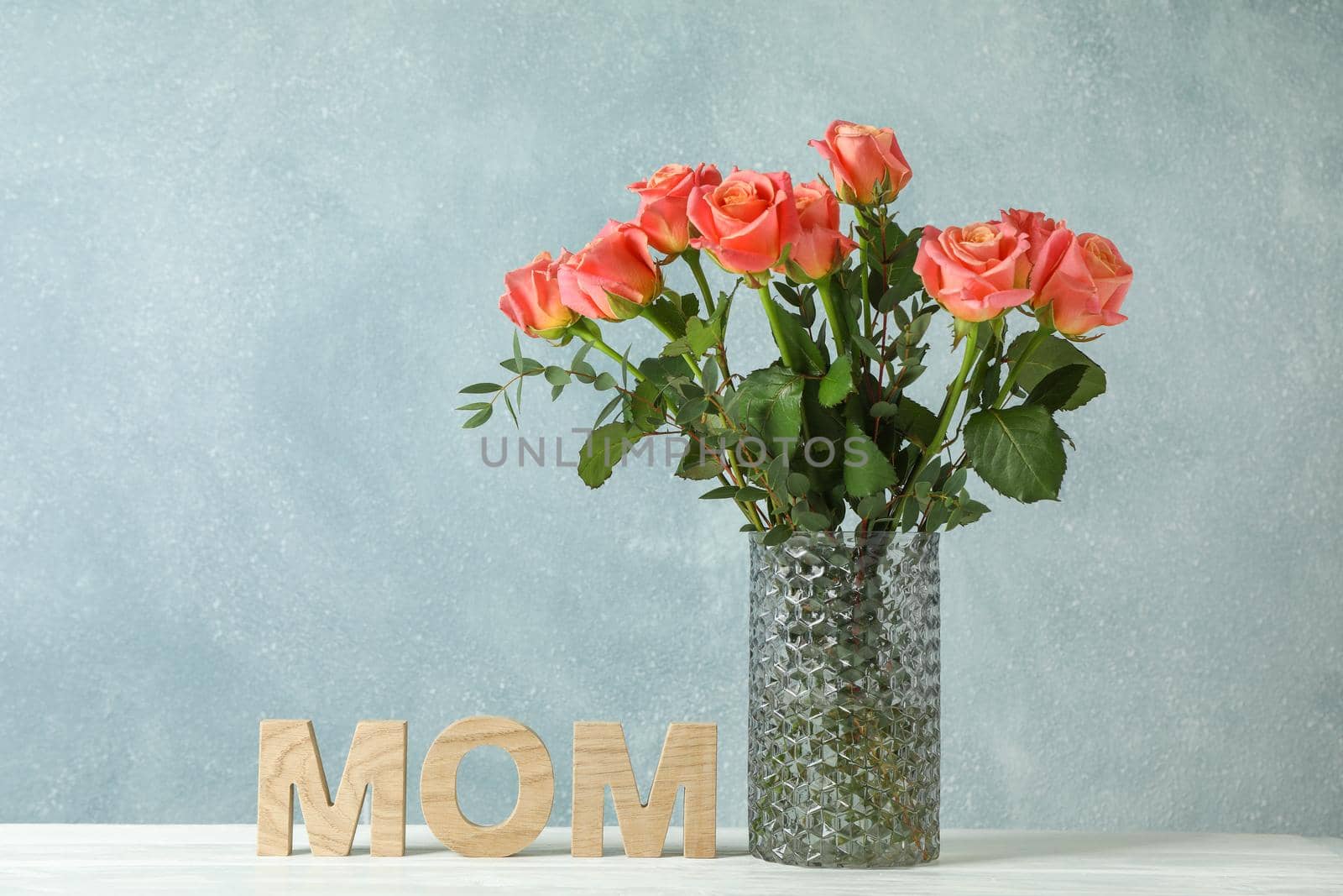 Vase with orange roses and inscription Mom on white table against light background, space for text