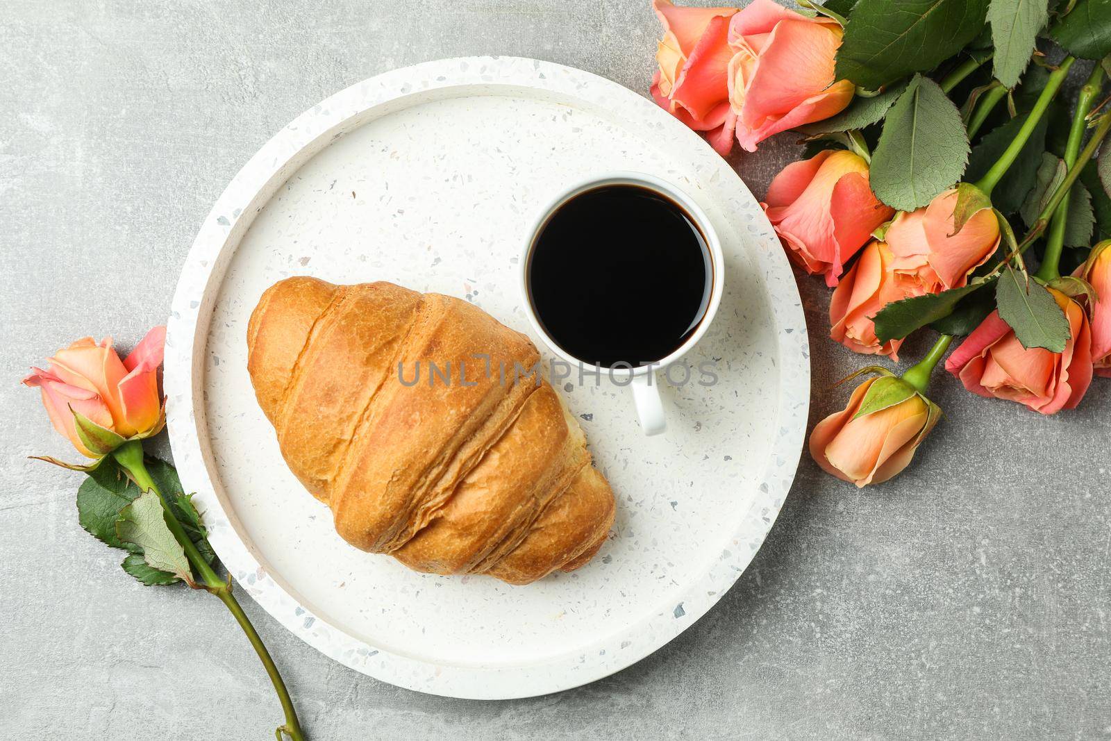Composition with marble tray, cup of coffee, croissant and roses on grey background, top view