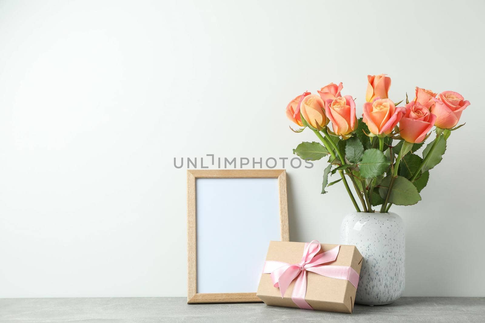 Vase with roses, gift and empty frame on grey table against white background, space for text