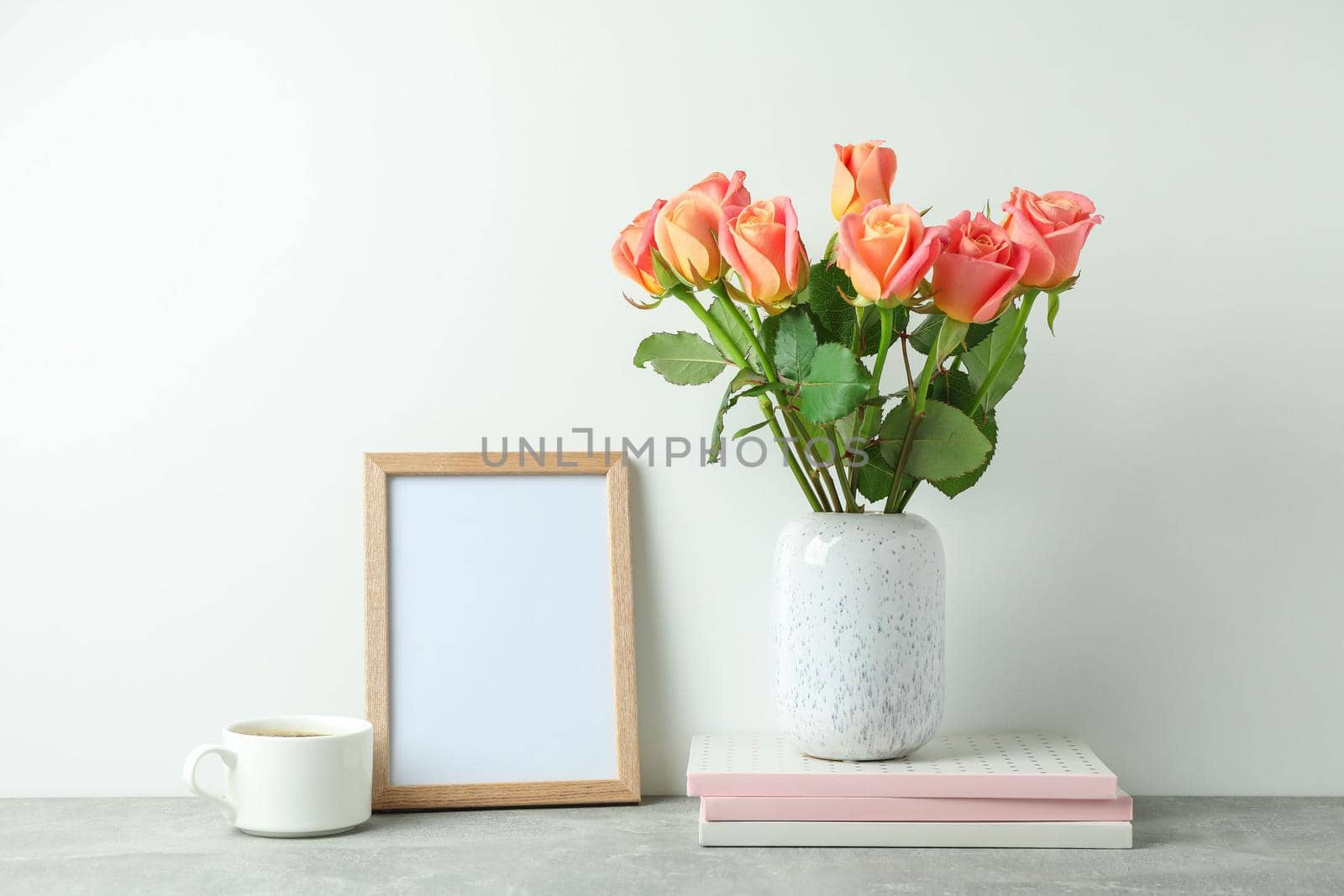 Vase with pink roses, copybooks, empty frame, cup of coffee on grey table against white background