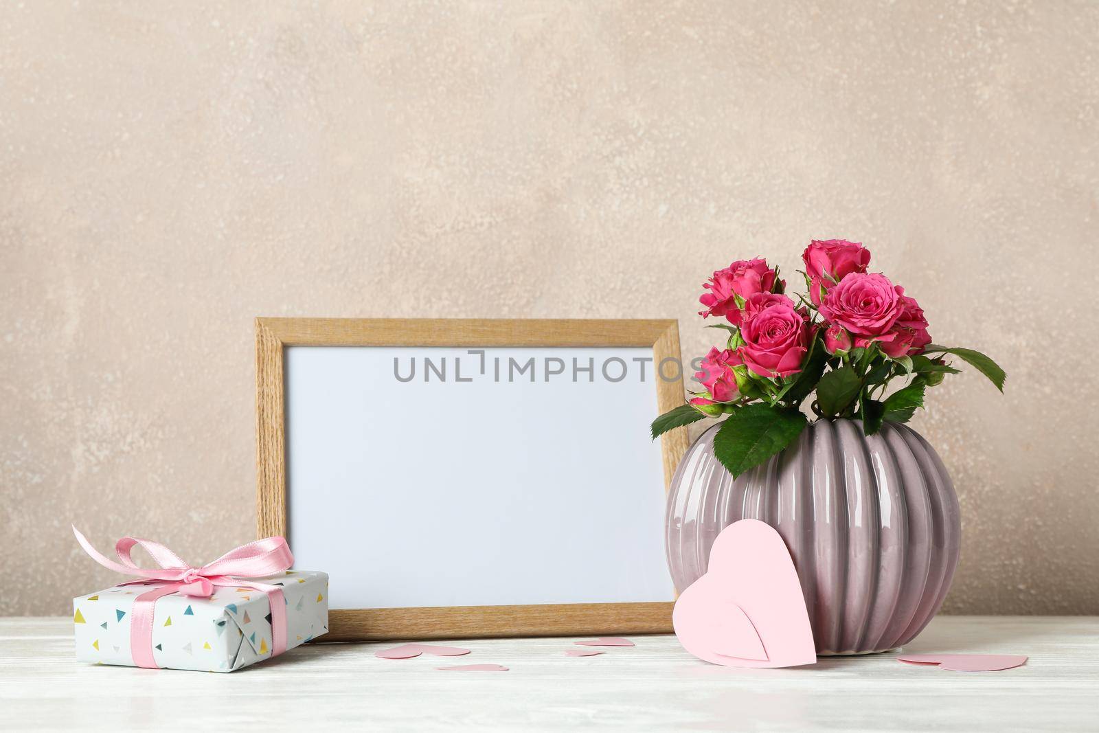 Vase with pink roses, empty frame, gift and little hearts on white table against light brown background by AtlasCompany