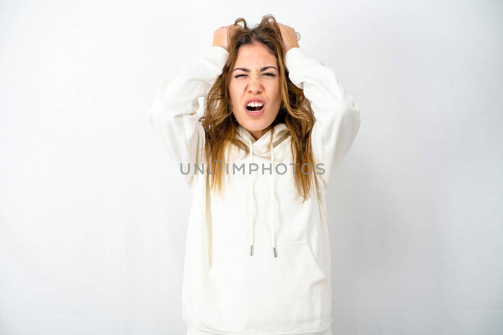 Young model isolated on white background tears hair out with despair face expressing anger by screaming with her mouth open and closed eyes. Concept of modern times stressed and frustrated woman