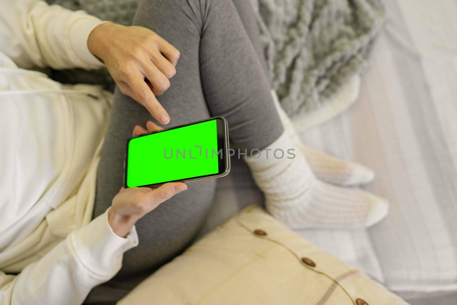 Bored girl sitting snuggled between the blankets and the sofa cushions pointing at her smartphone with green screen. New nodern times home habit to wasting time with the internet and social networks by robbyfontanesi