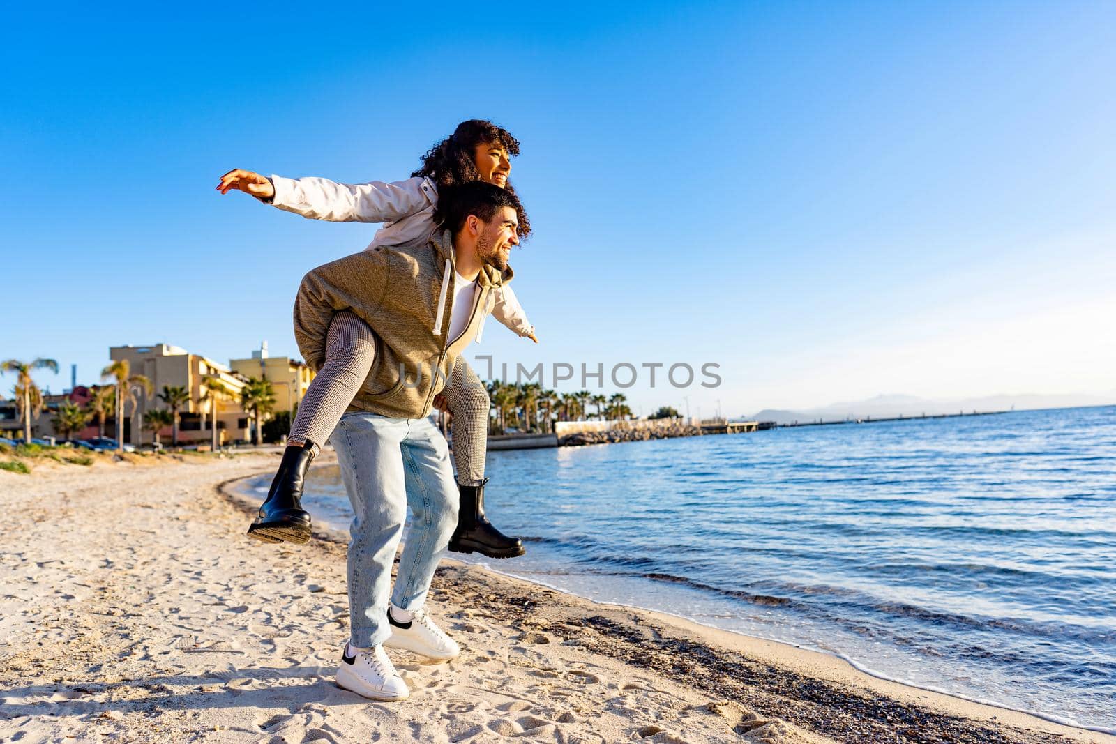 Happy multiracial young beautiful couple standing on the beach smiling at the setting sun on the sea or ocean water. Afro american curly beautiful girl imitates plane wings in piggyback on boyfriend by robbyfontanesi