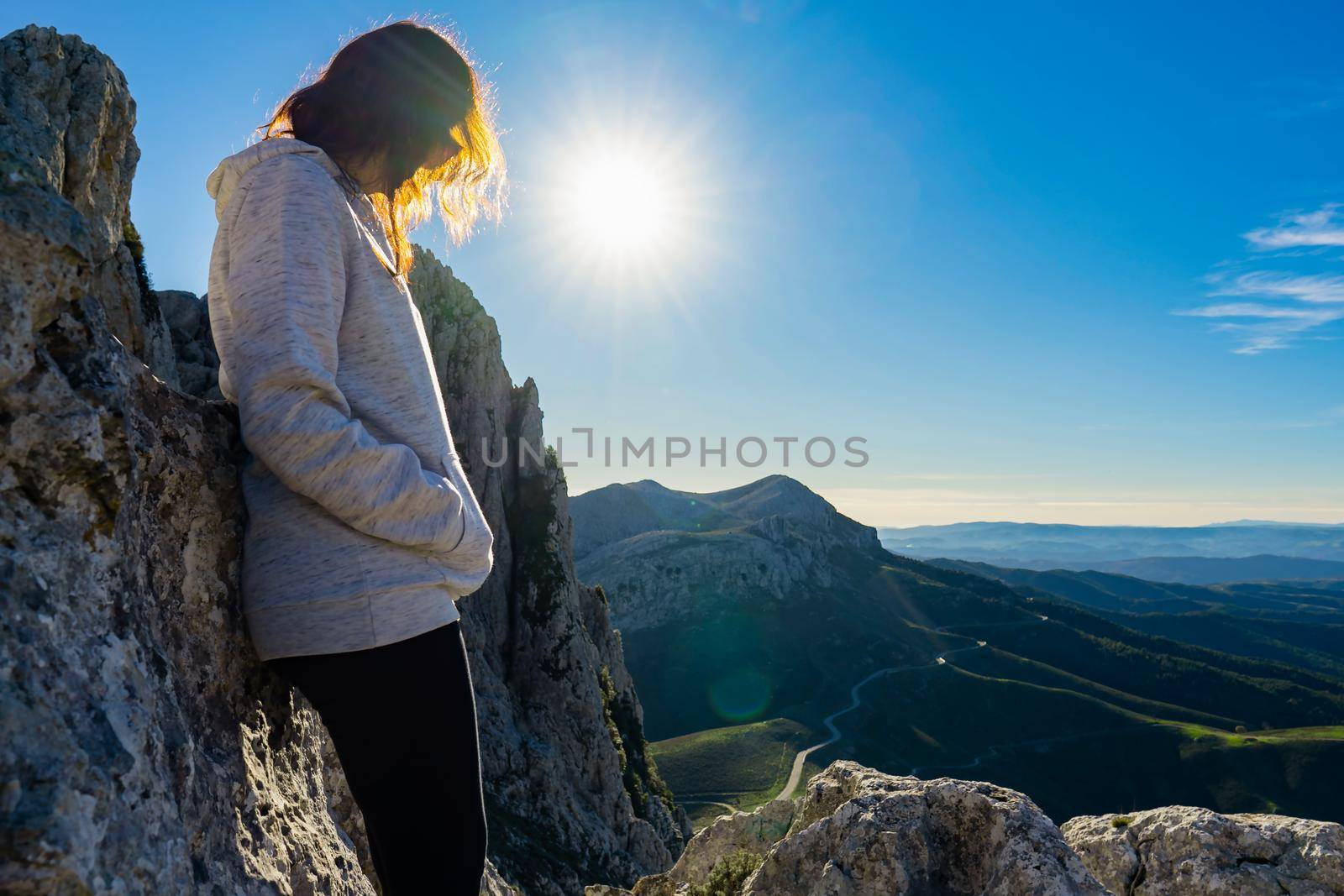 Lonely woman finds herself leaning against the rocks in the nature pensive at top of mountains with a splendid view below and sun through her hair. Concept of positive loneliness to find your own way