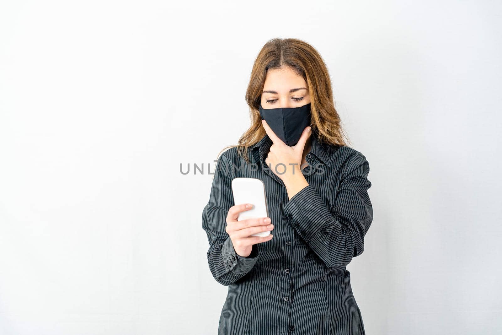 Young model woman isolated on white background wearing business dark shirt and protective mask keeps her chin worried looking smartphone reading latest Coronavirus news. Covid girl expressions by robbyfontanesi