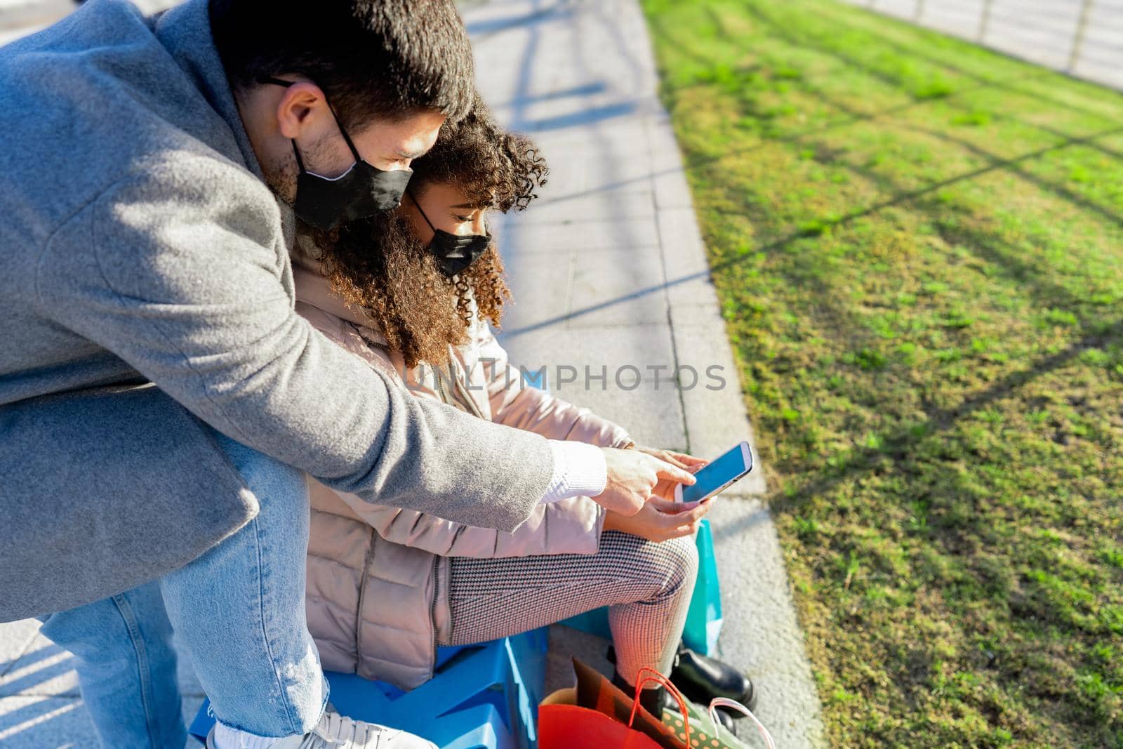 Young couple in Coronavirus protective mask use smartphone to shop online outdoors. Afro american girl sitting on a city park bench using internet with her boyfriend for online shopping