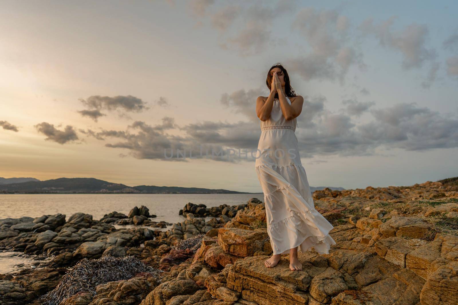Pensive Woman in long white dress on with folded hands in front of face and closed eyes contemplating the sunset over the sea. Concept of experiencing nature to feel good and find your spirituality