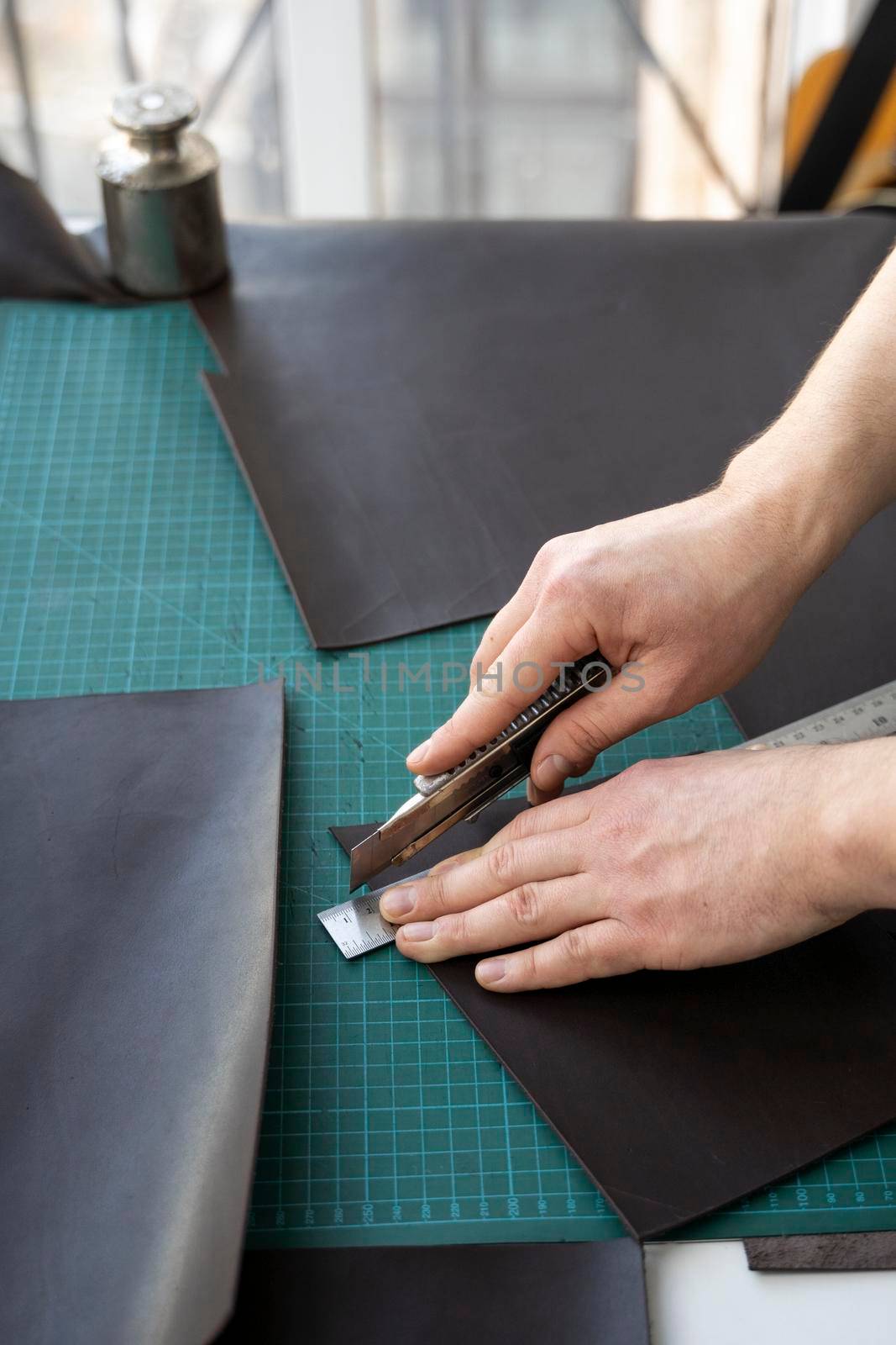 Men's hand holding a stationery knife and metal ruler and cutting on a pieces for a leather wallet in his workshop. Working process with a brown natural leather. Craftsman holding a crafting tools