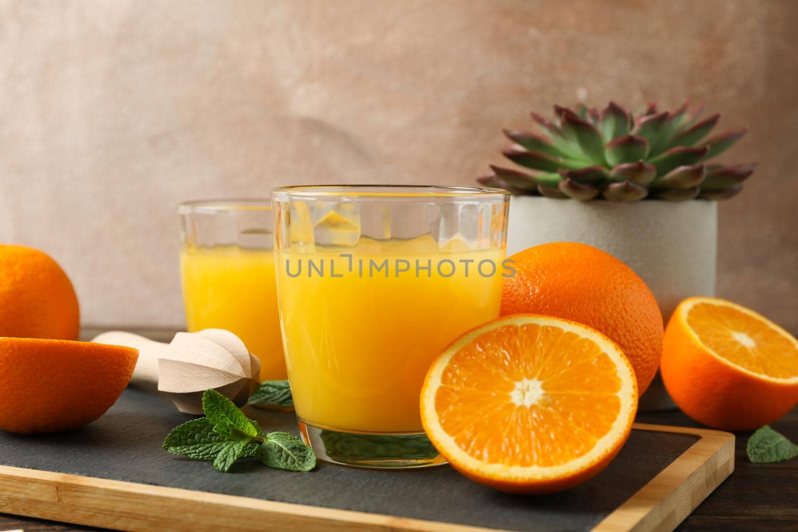 Cutting board, orange juice, wooden juicer, mint, orange, tubule and succulent plant on wooden table, space for text. Fresh natural drink and fruits