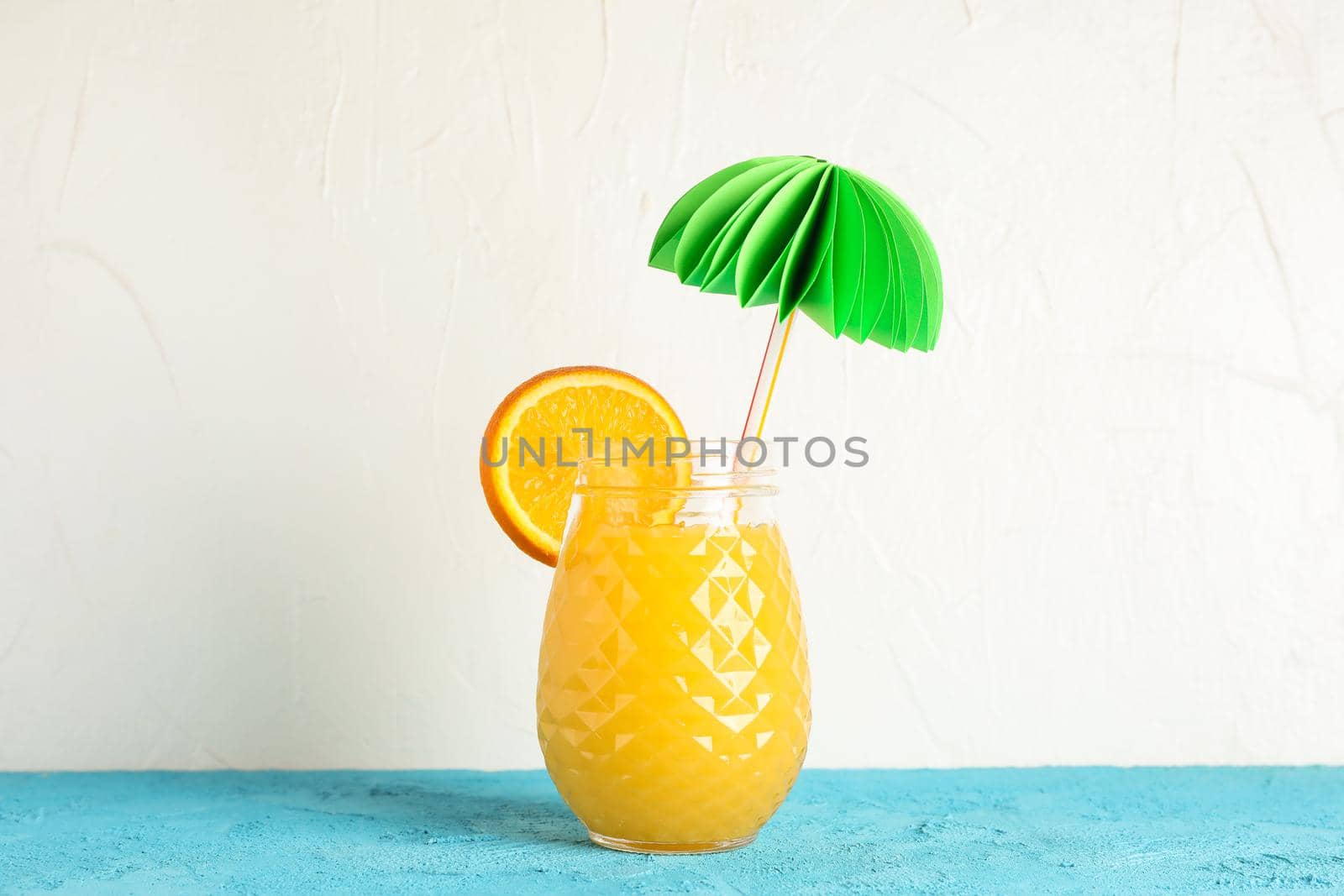 Glass jar with decorative palm and orange piece on color table against white background, space for text. Fresh natural drink by AtlasCompany