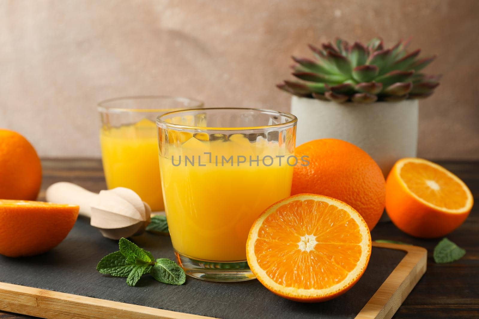 Cutting board, orange juice, wooden juicer, mint, orange, tubule and succulent plant on wooden table, space for text. Fresh natural drink and fruits