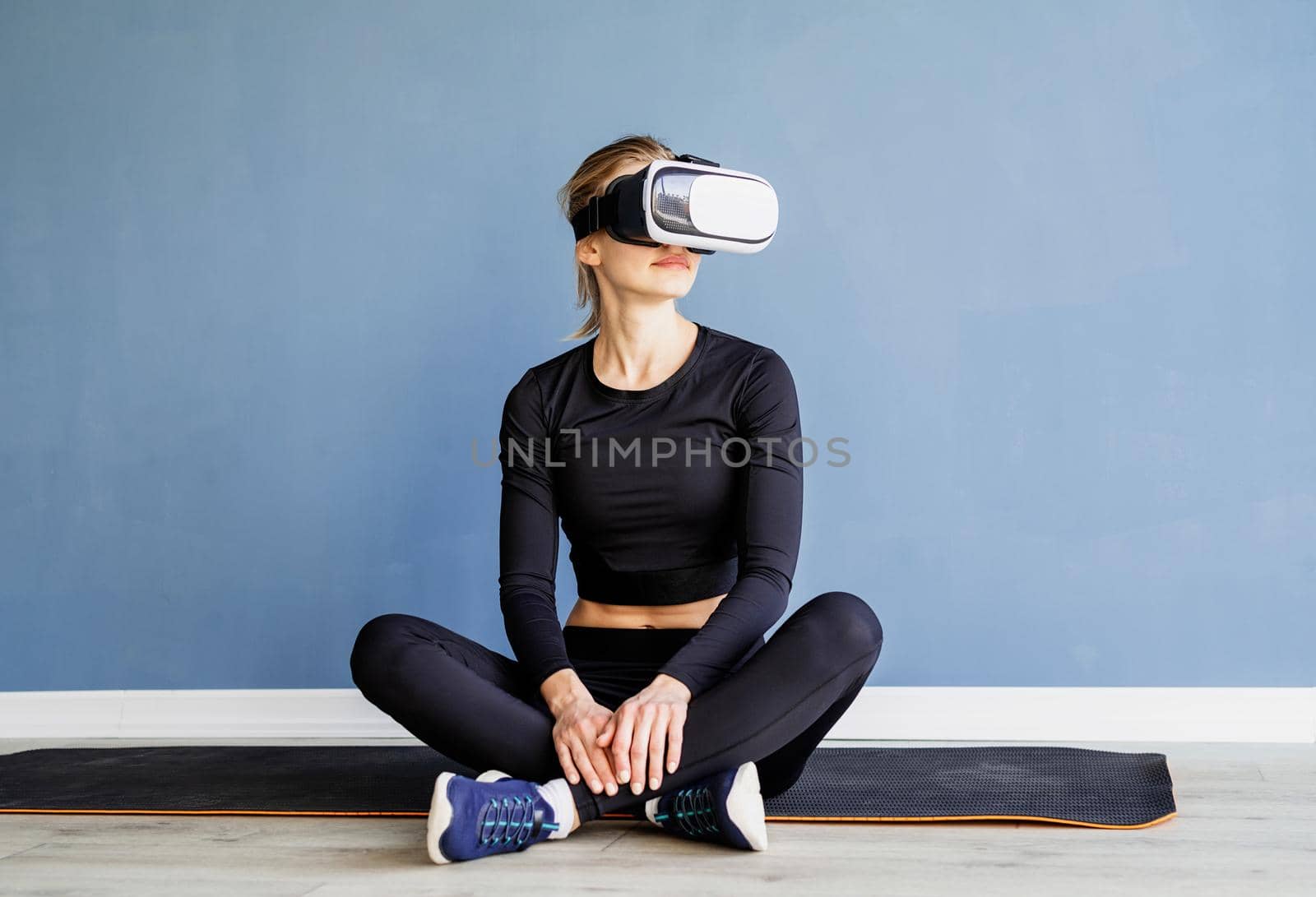 Young blonde woman in sport clothes wearing virtual reality glasses sitting at fitness mat at at home by Desperada