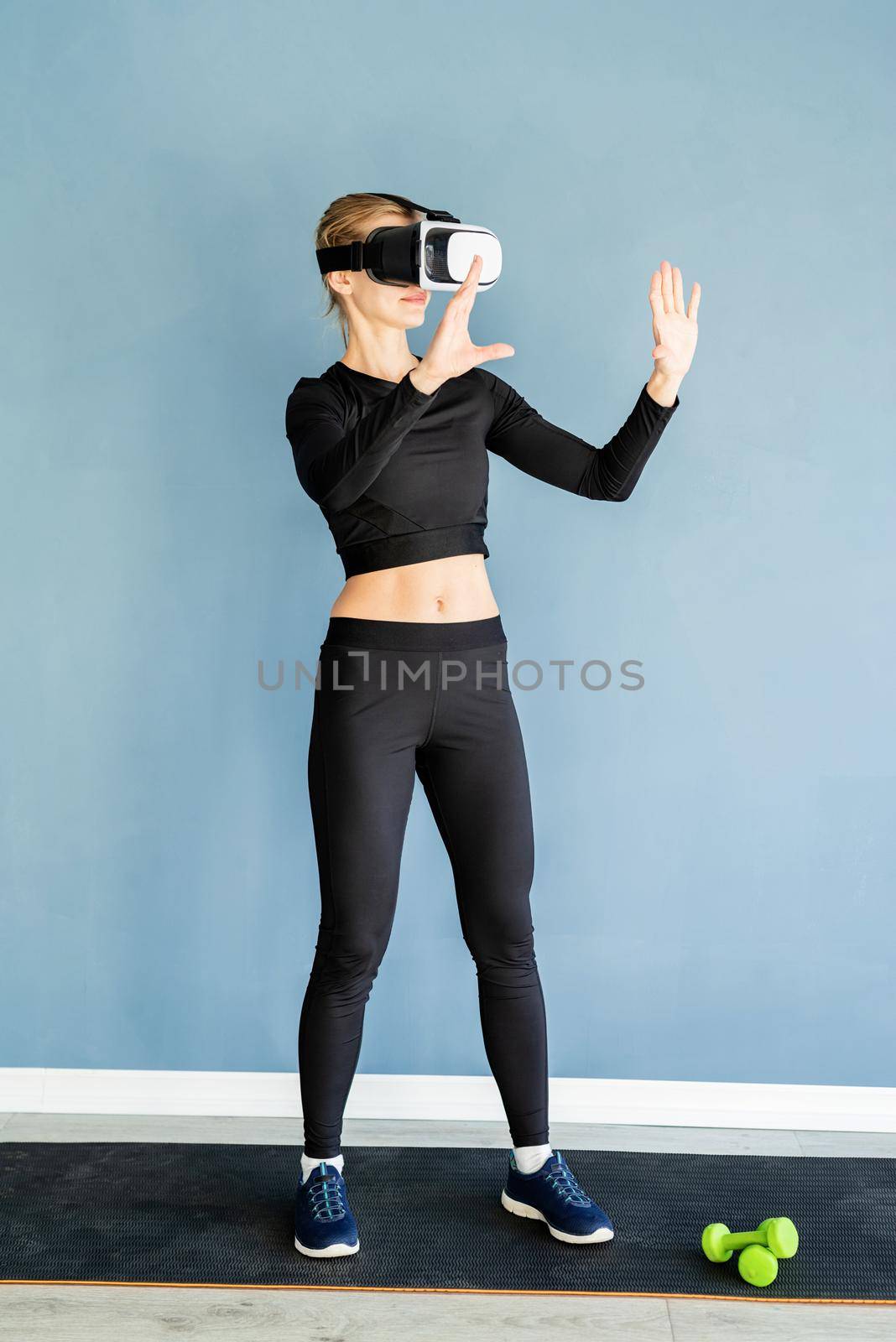 Young blonde woman in sport clothes wearing virtual reality glasses standing at fitness mat using VR interactive menu by Desperada
