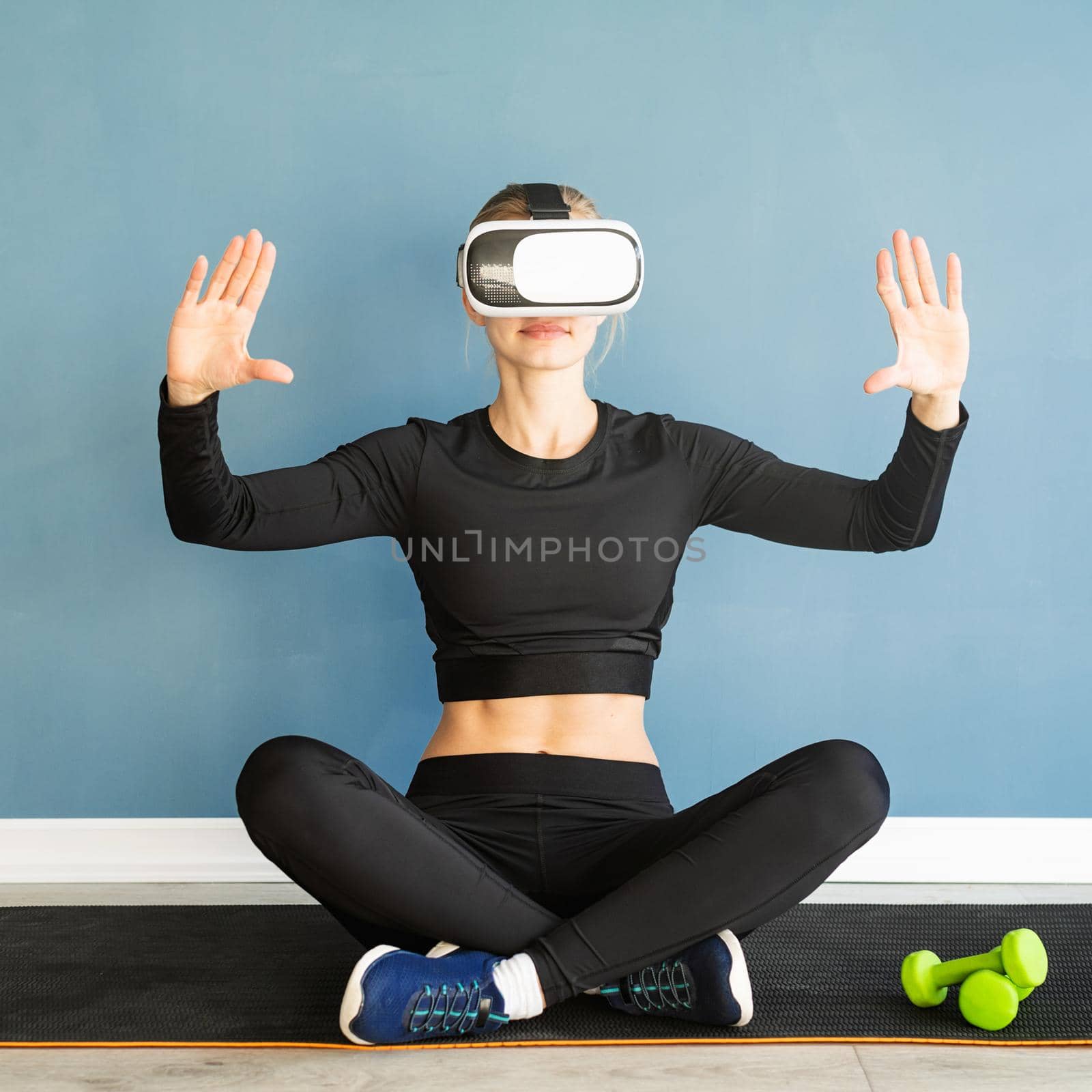 Young blonde woman in sport clothes wearing virtual reality glasses sitting at fitness mat using VR interactive menu by Desperada