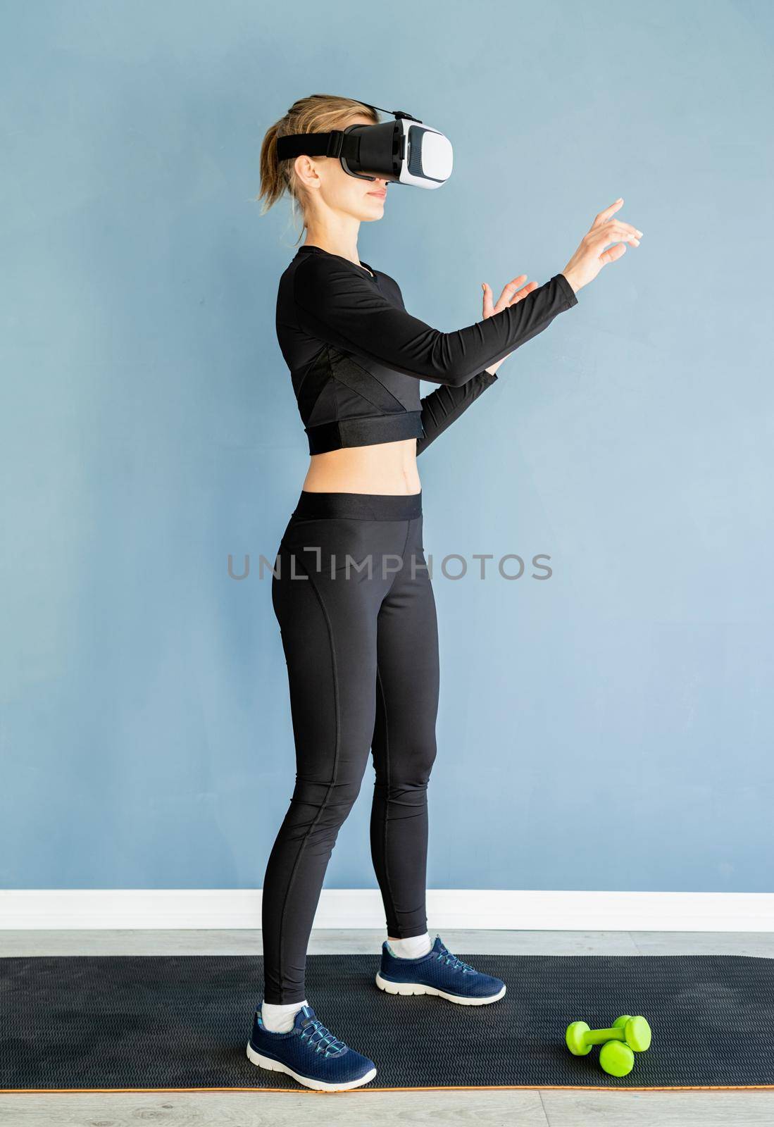 Young blonde woman in sport clothes wearing virtual reality glasses standing at fitness mat using VR interactive menu by Desperada