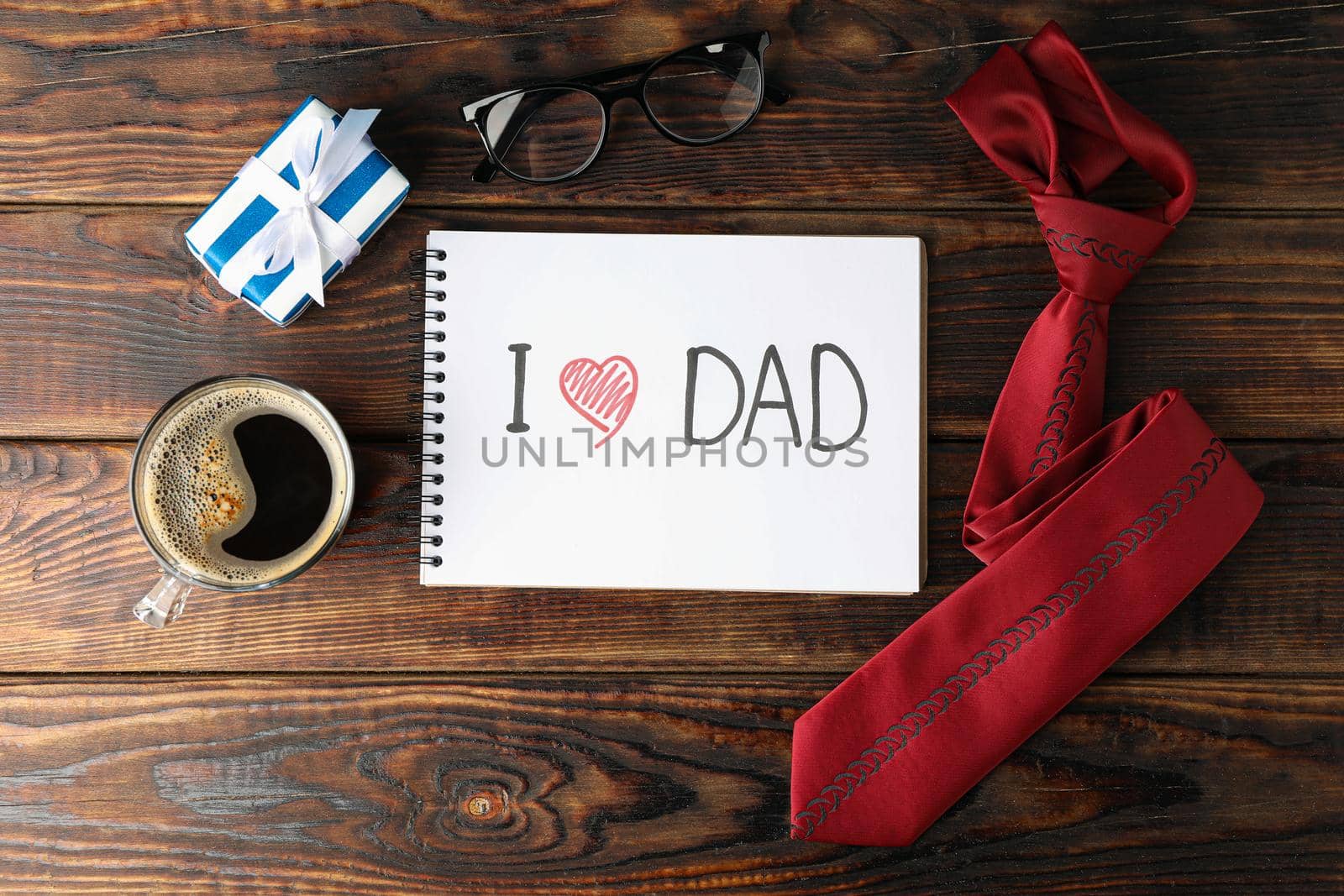 Notebook with inscription I love DAD, cup of coffee, glasses, gift box and tie on wooden background, space for text and top view