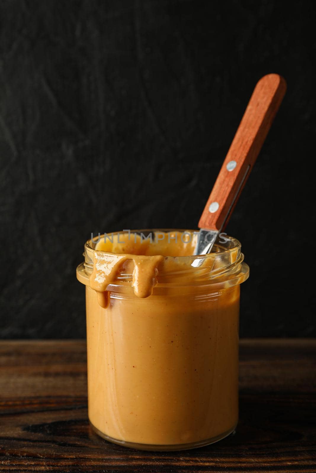 Glass jar with peanut butter and spoon on wooden table against black background, space for text