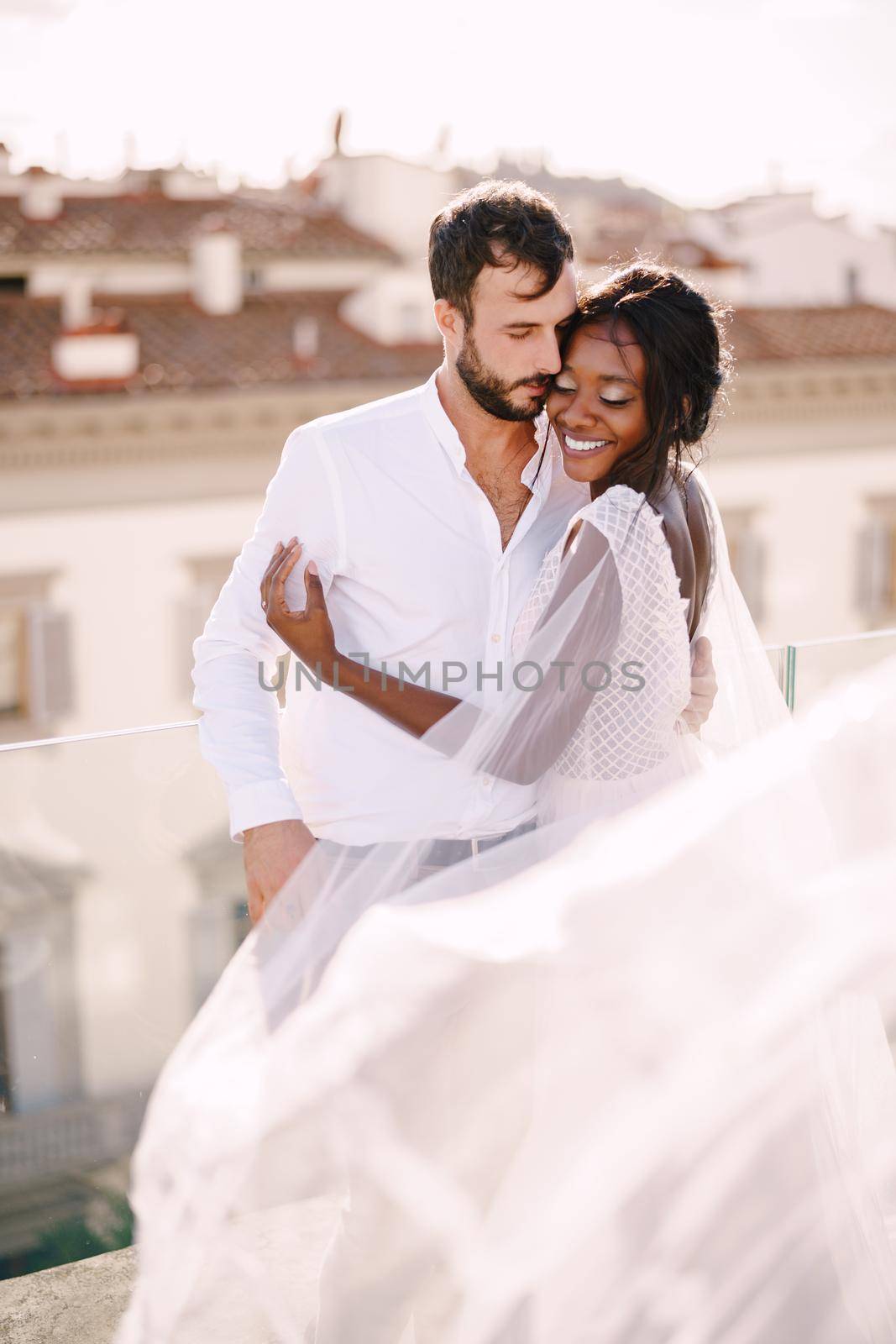 Destination fine-art wedding in Florence, Italy. Multiethnic wedding couple. African-American bride hugs Caucasian groom on the roof with cityscape view. by Nadtochiy