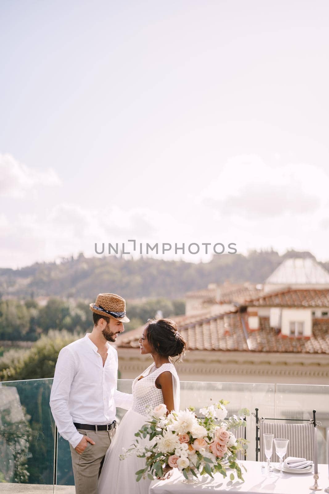 Destination fine-art wedding in Florence, Italy. Multiethnic wedding couple. African-American bride and Caucasian groom with a beard and straw hat, near the table for a wedding dinner. by Nadtochiy