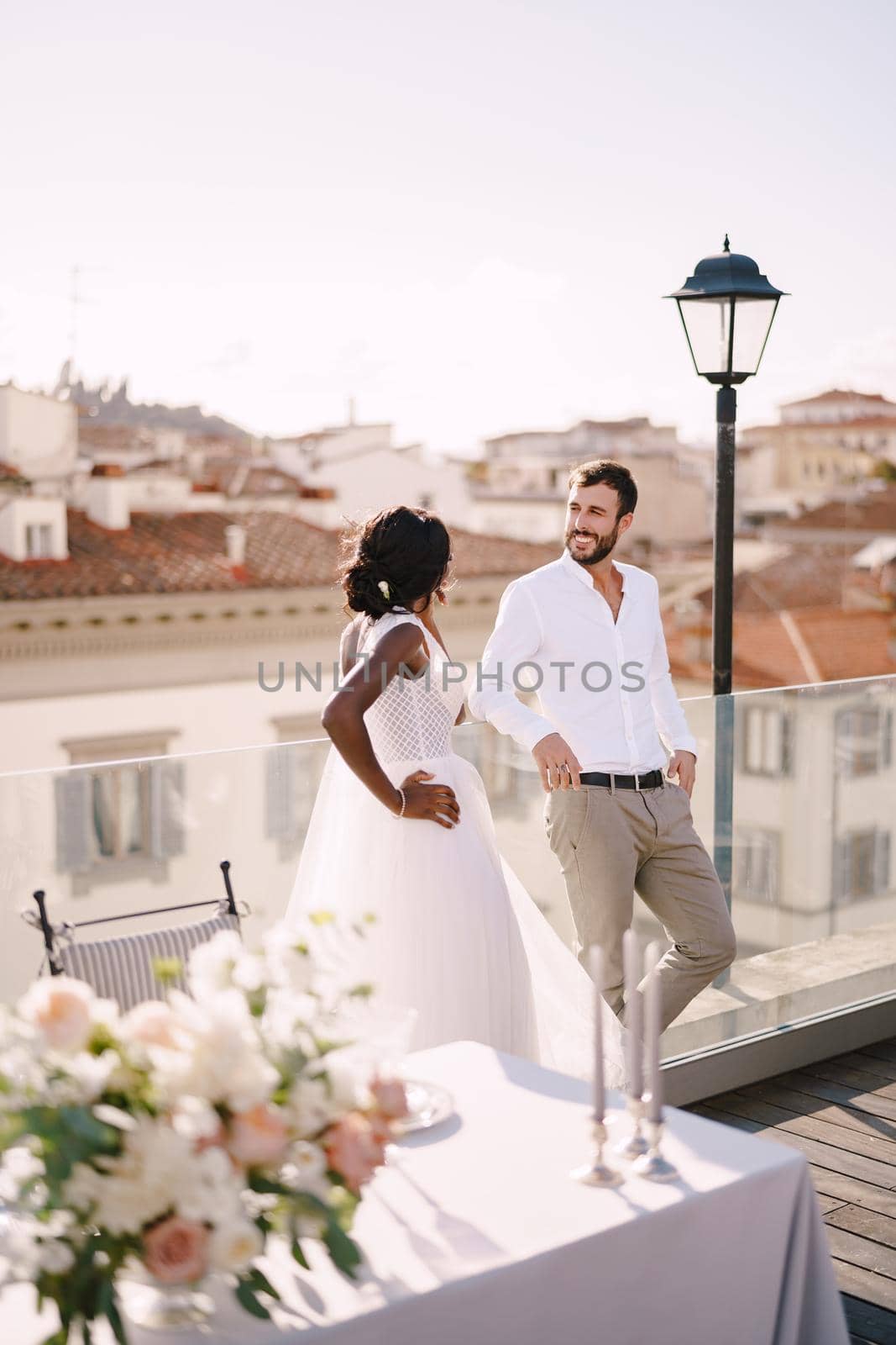 Multiracial wedding couple. Destination fine-art wedding in Florence, Italy. African-American bride and Caucasian groom stand near the table for a wedding dinner. by Nadtochiy