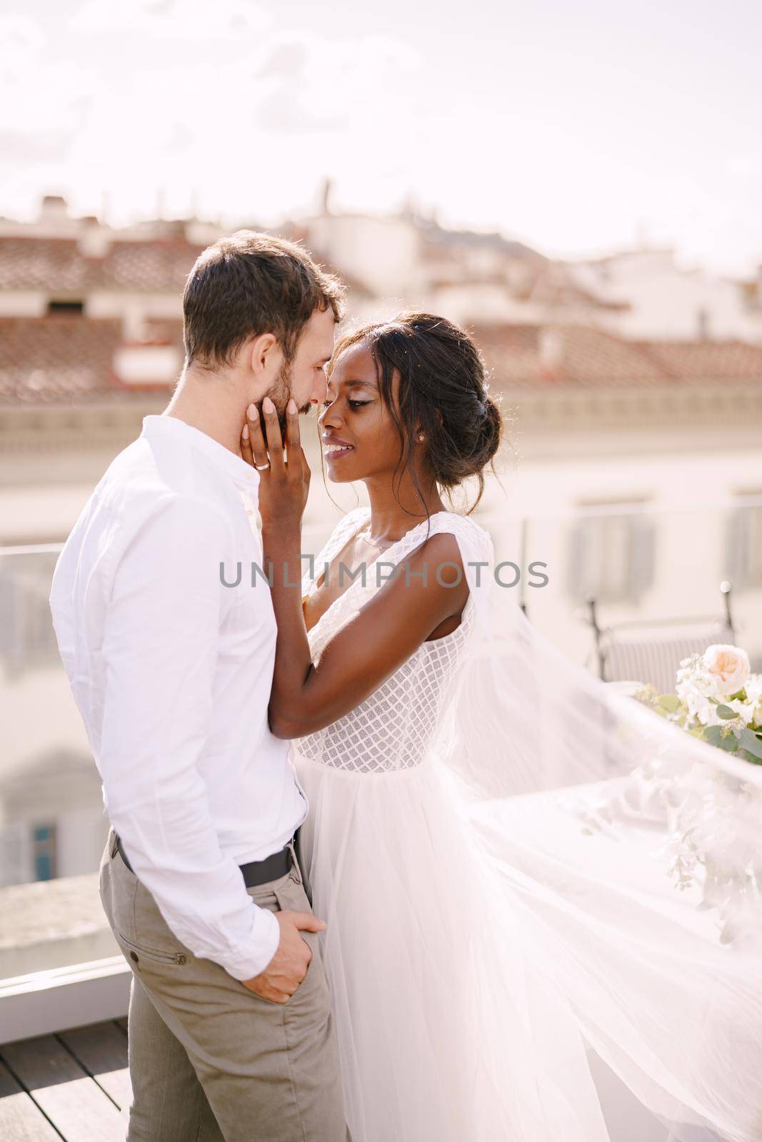 Mixed-race wedding couple. Caucasian groom and African-American bride cuddling on a rooftop in sunset sunlight. Destination fine-art wedding in Florence, Italy. by Nadtochiy