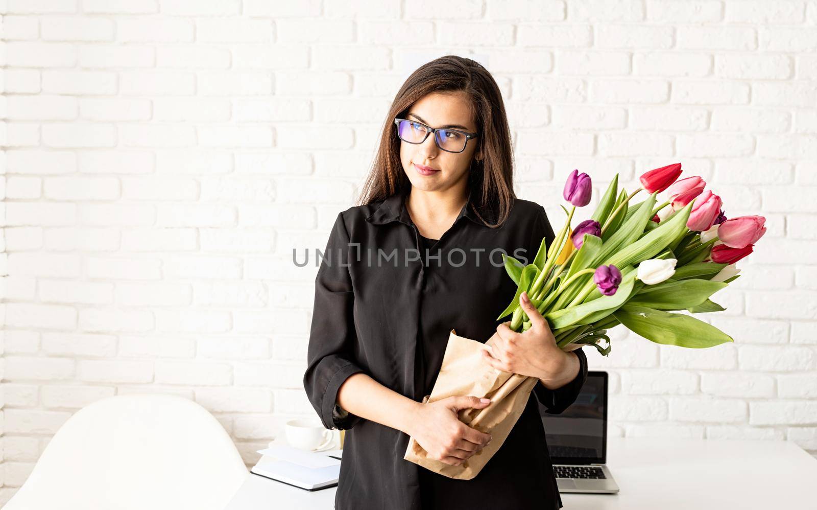 Portrait of young confident brunette business woman standing by the desk with fresh tulips bouquet by Desperada