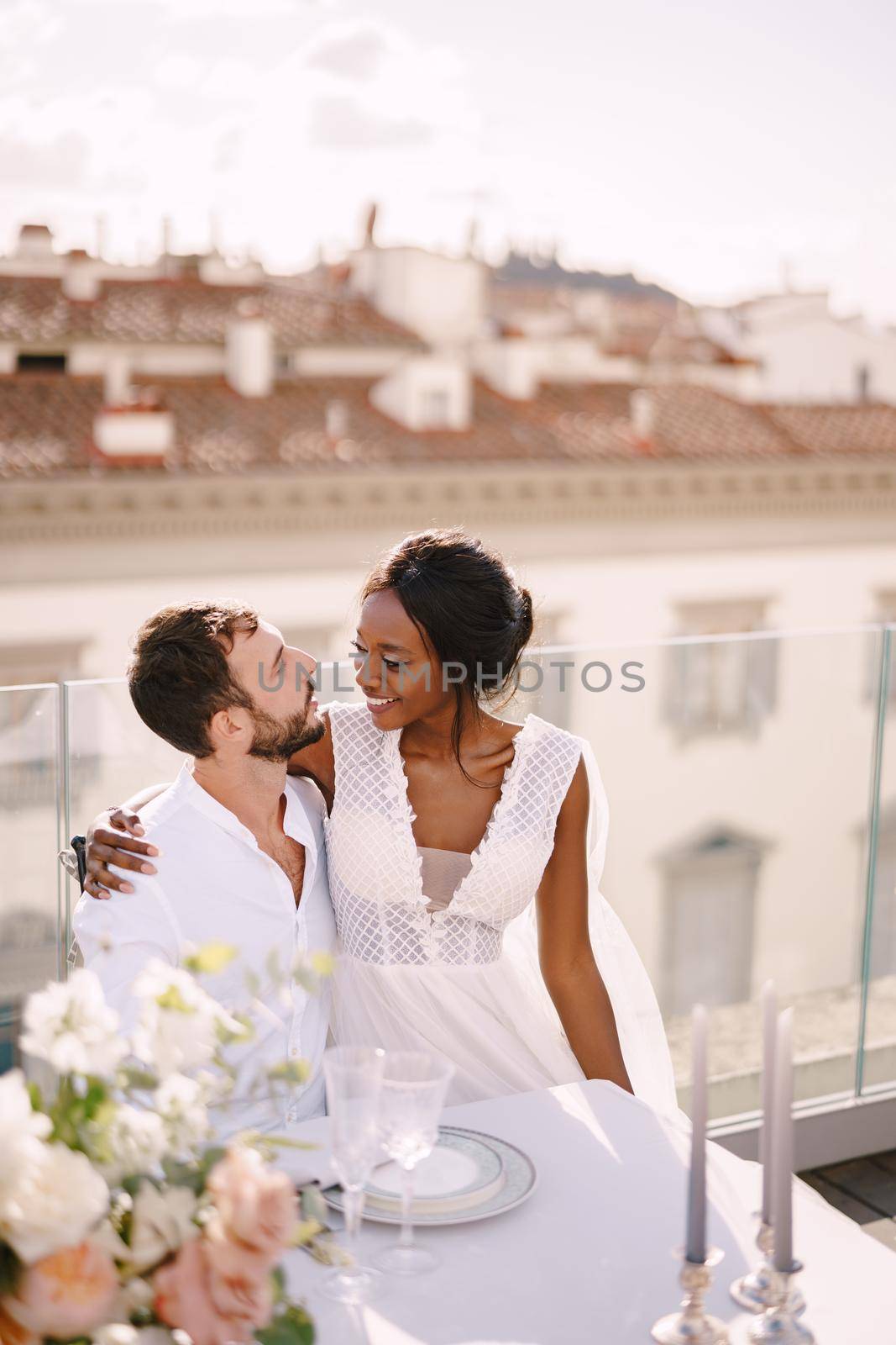 Destination fine-art wedding in Florence, Italy. Multiethnic wedding couple. African-American bride and Caucasian groom are sitting at the rooftop wedding dinner table overlooking the city. by Nadtochiy