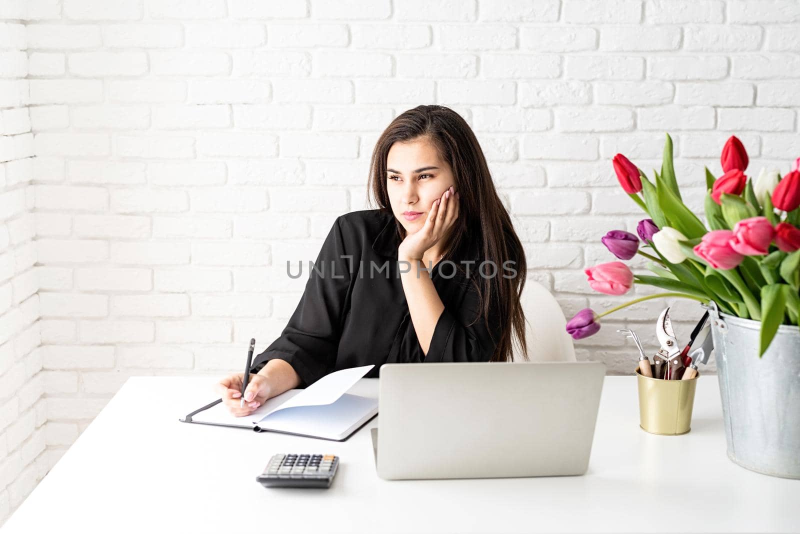 A young business woman florist writing in notebook, using laptop in the office, bucket of tulips on the desk. Small business.