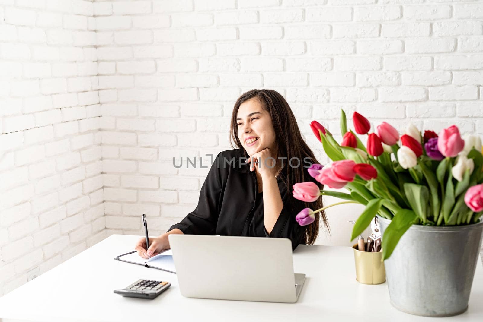 young brunette business woman florist writing in notebook, using laptop in the office, bucket of tulips on the desk. Small business.