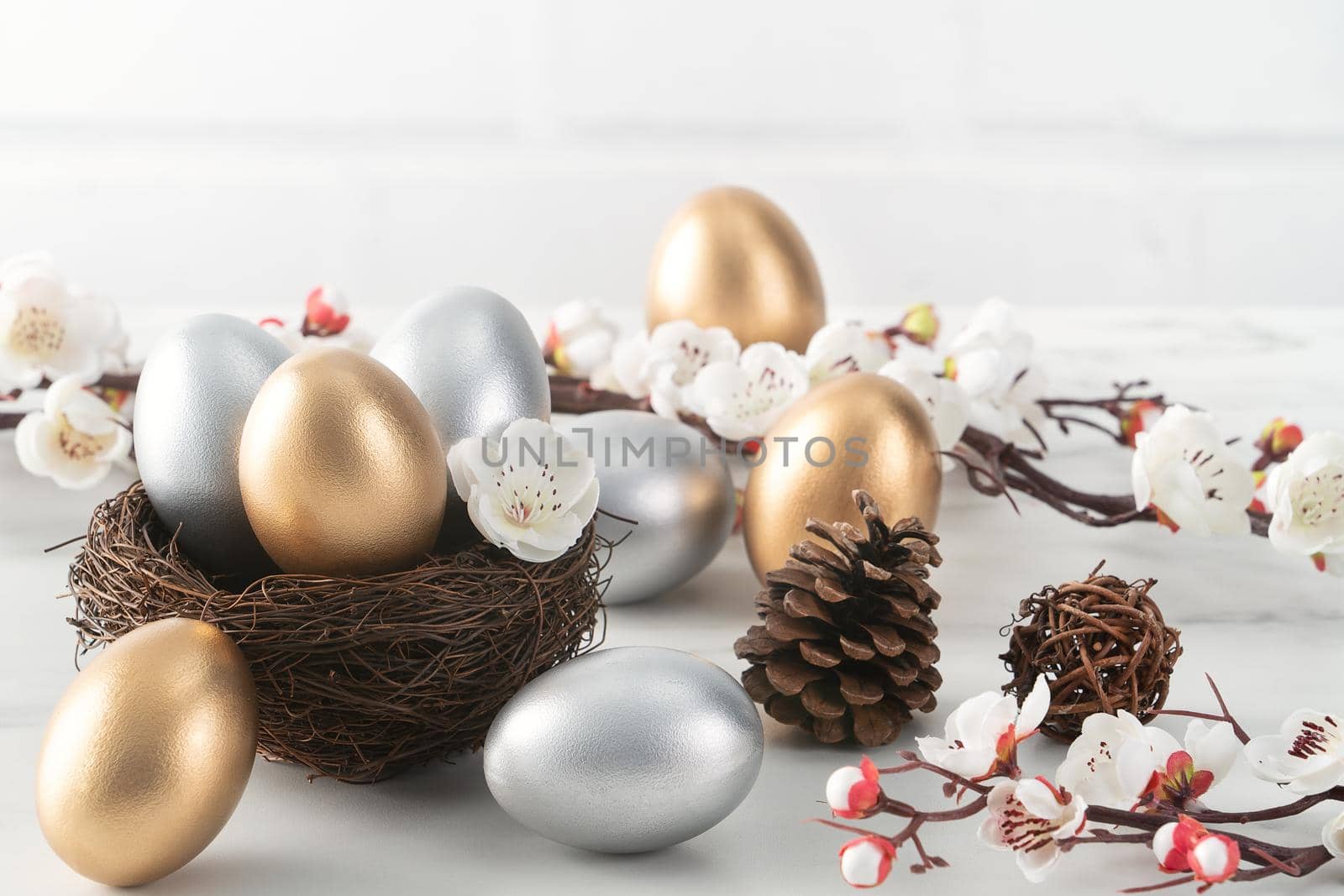 Close up of golden and silver Easter eggs in the nest with white plum flower on bright white wooden table background.