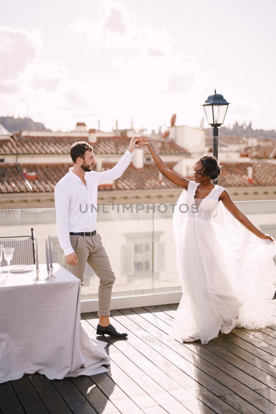 Interracial wedding couple. Destination fine-art wedding in Florence, Italy. African-American bride and Caucasian groom are dancing near the table for a wedding dinner, on the roof of the restaurant. by Nadtochiy