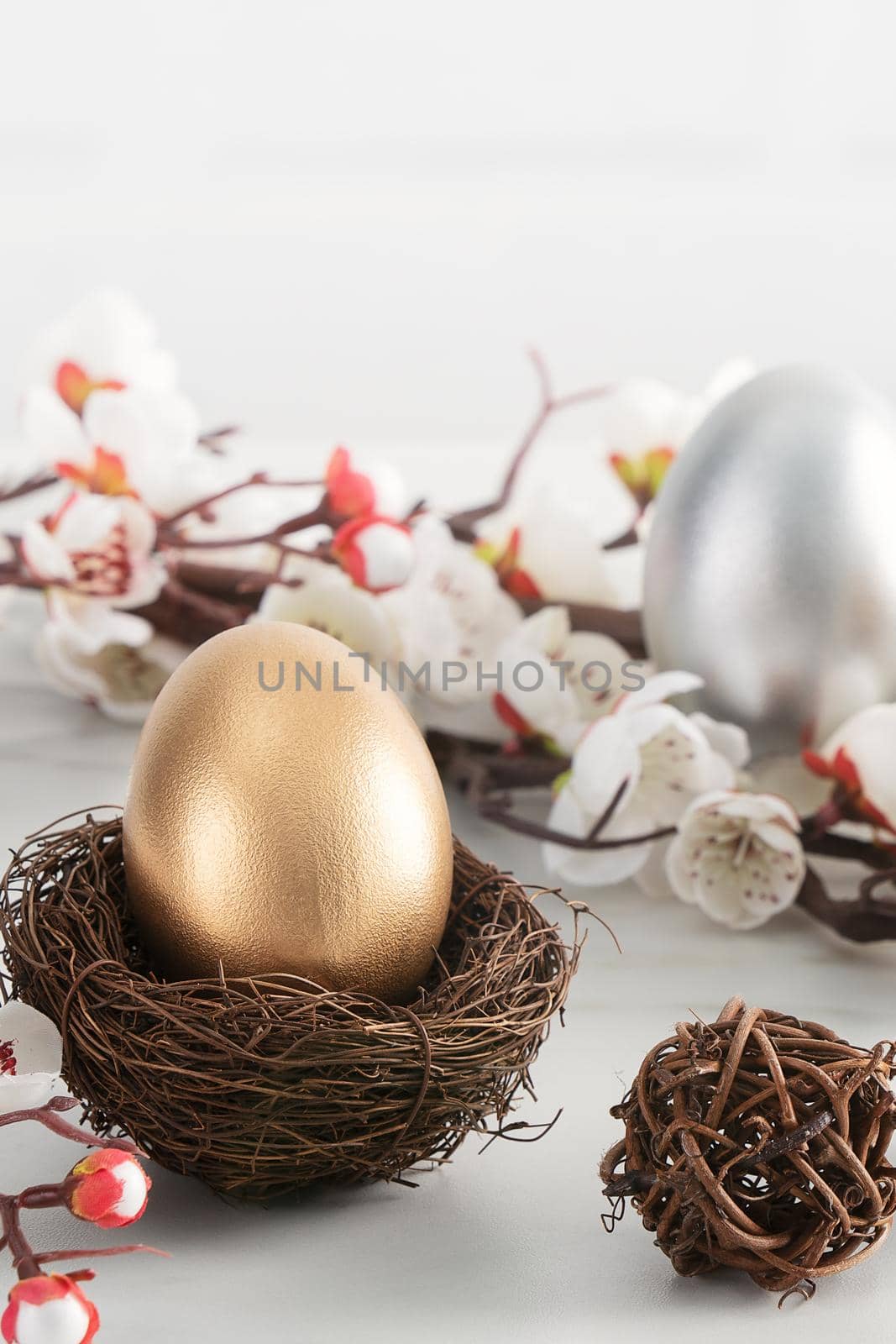 Close up of golden and silver Easter eggs in the nest with white plum flower on bright white wooden table background.